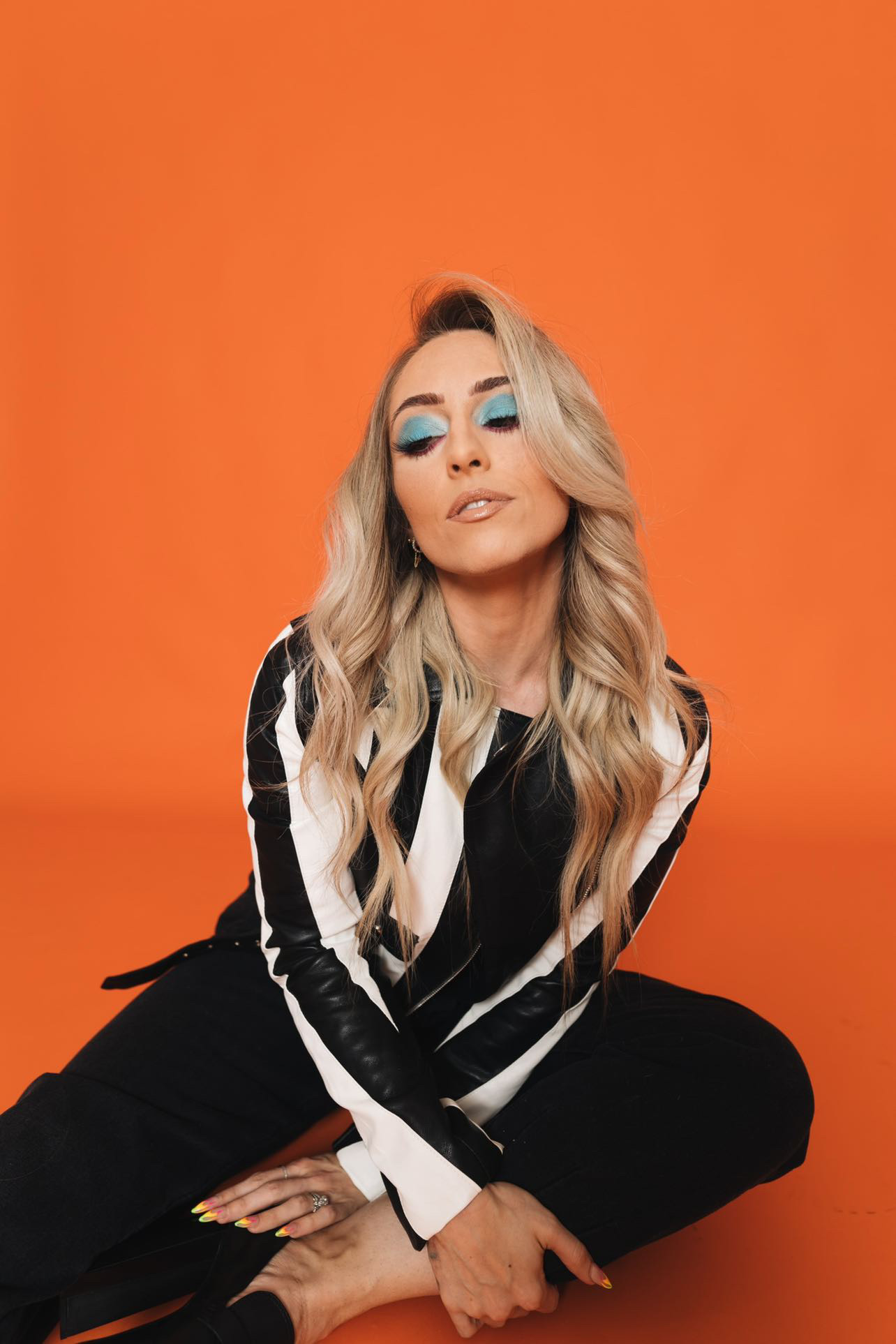 Woman with long blonde hair and blue eyeshadow sitting against an orange background.