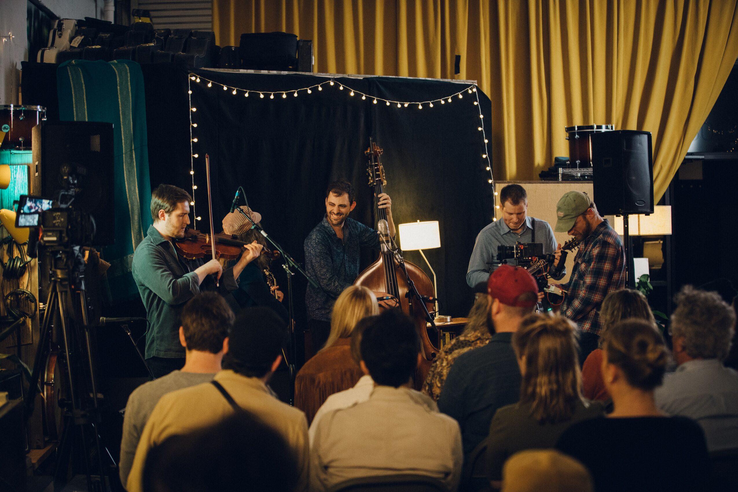 Musicians perform on stage with an audience watching, decorated with string lights and yellow curtains in the background.