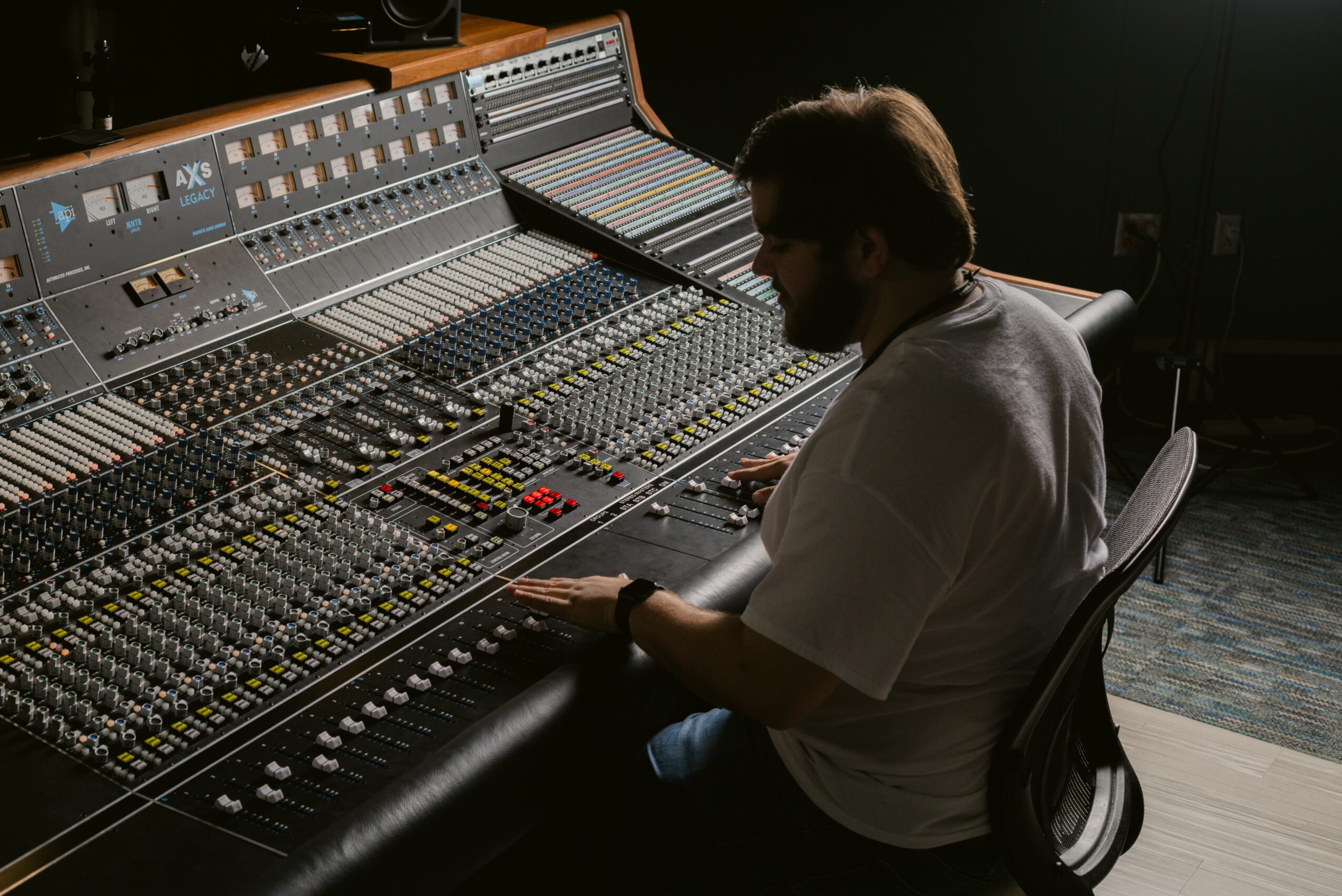 Person sitting at a large audio mixing console in a recording studio, working with controls and sliders.