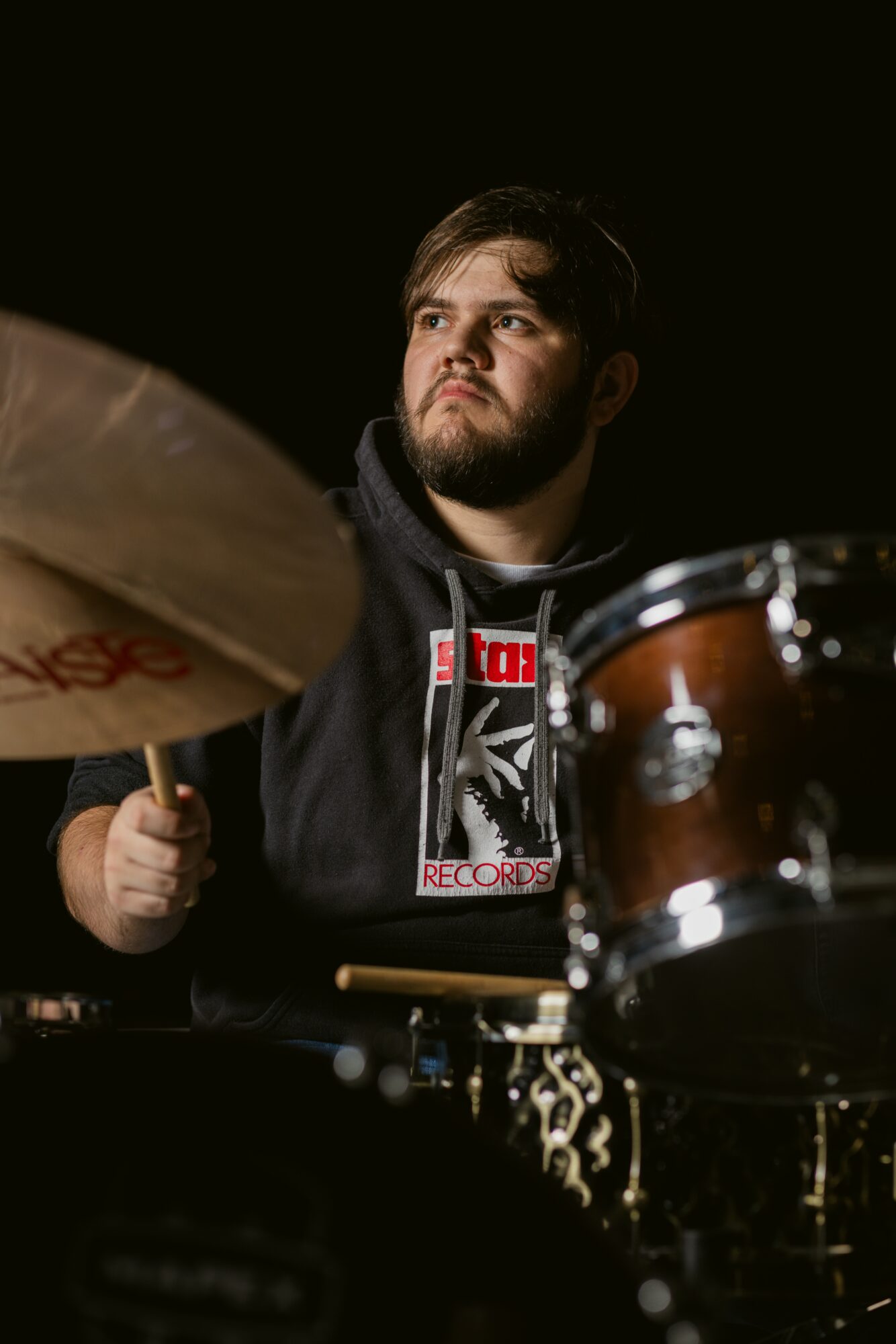 Young man with a beard playing drums, looking to the side, dark background, wearing a hoodie with a graphic design.