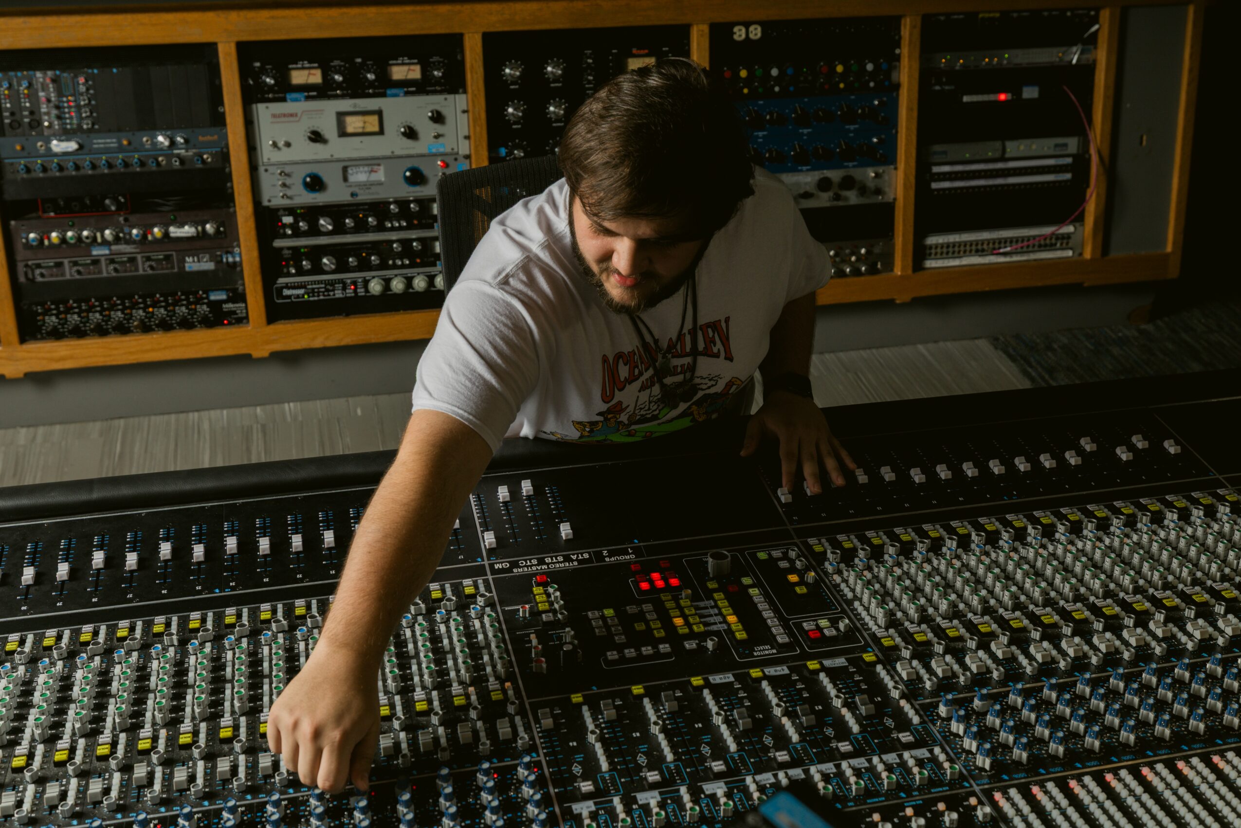 Person adjusting sound mixing console in a recording studio with audio equipment on shelves behind.