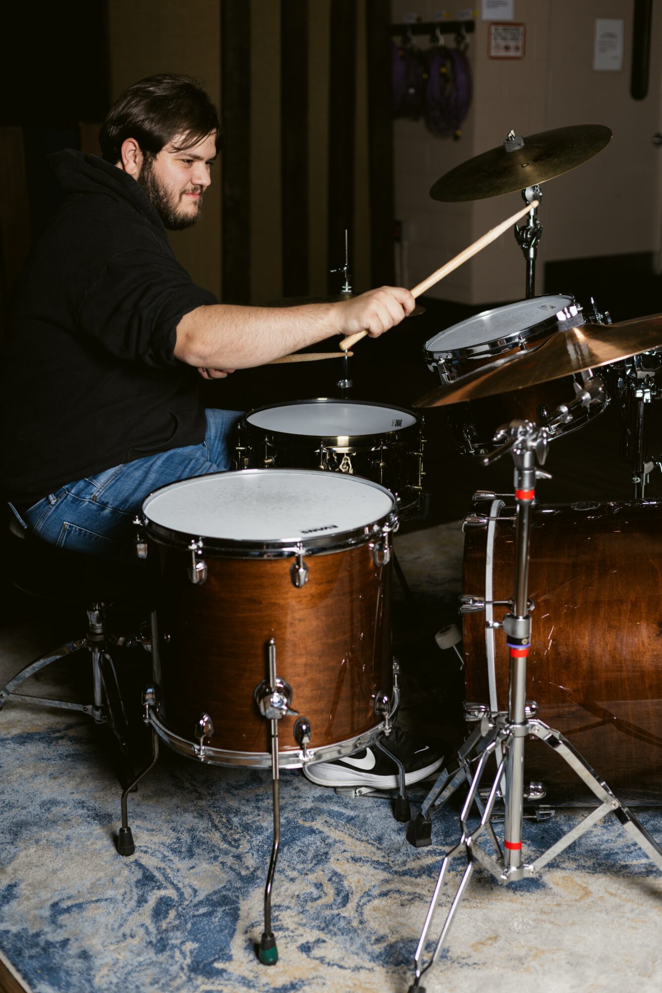 Person playing drums indoors, holding drumsticks, with drums and cymbals visible, dark background, patterned floor.