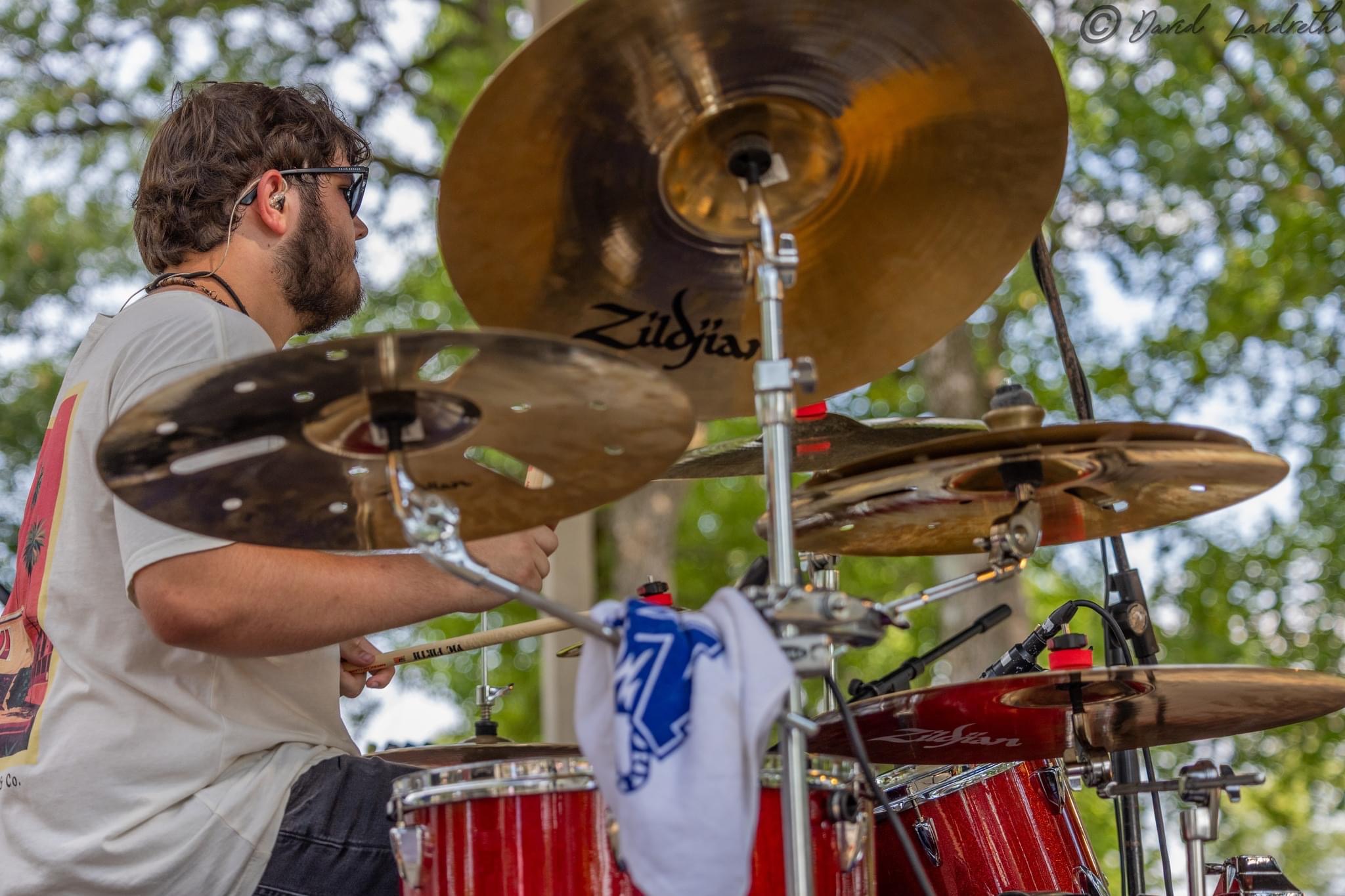 A man with glasses plays a drum set outdoors, with cymbals and drums visible, trees in the background.