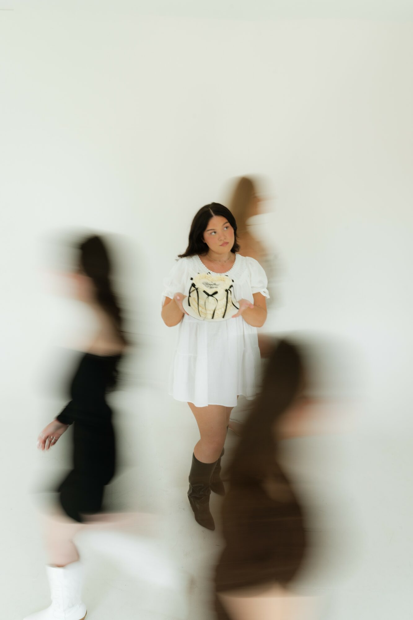 A woman in a white dress with a graphic print stands against a plain background, surrounded by blurred figures.