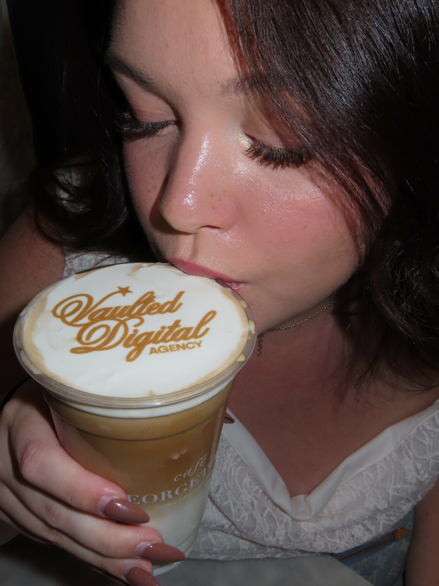 Woman with dark hair holding a glass with a logo, close-up of face and drink, in a dim setting.