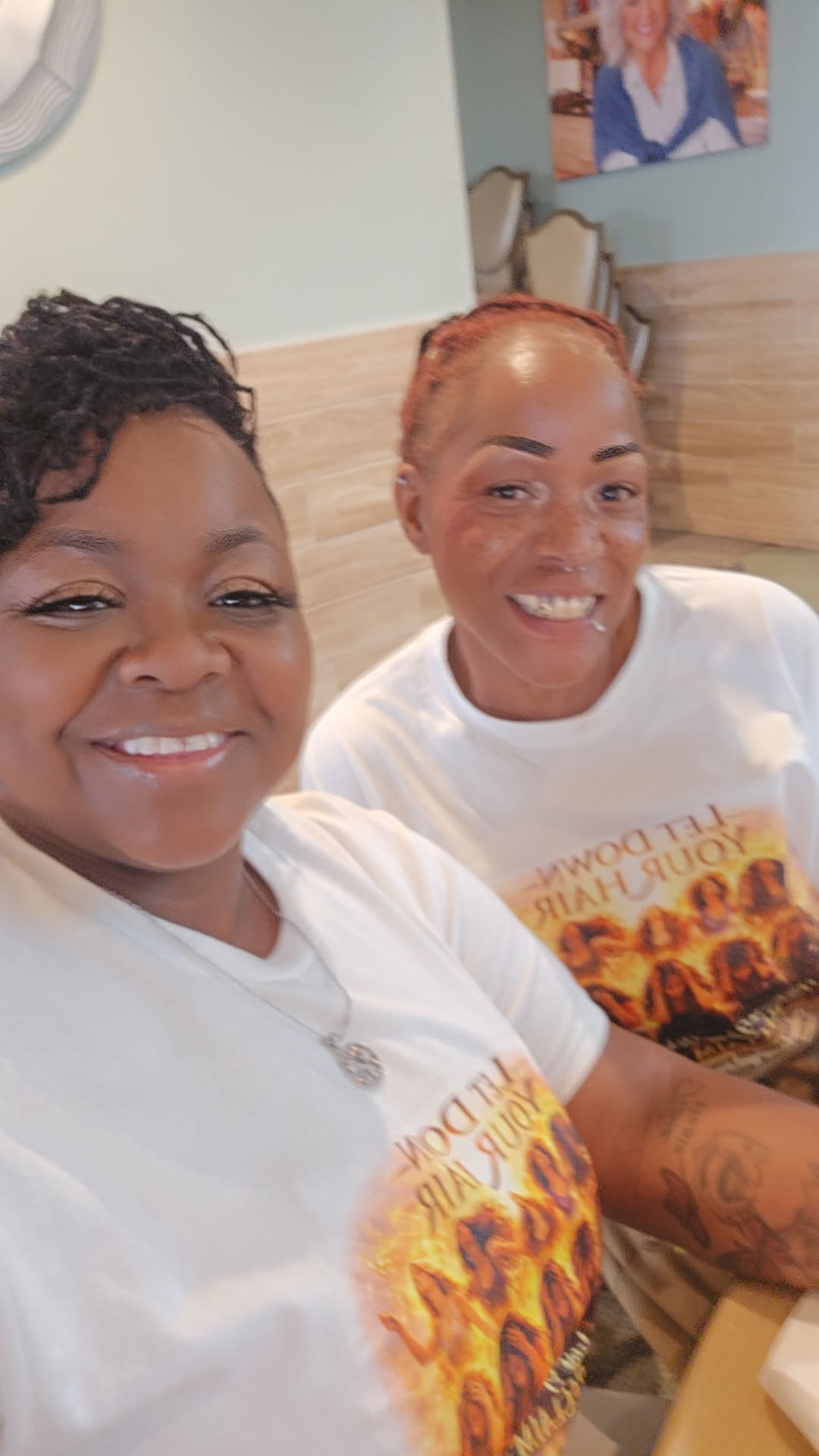 Two smiling women wearing matching white shirts with colorful text, sitting indoors with a light-colored wall and framed photo behind them.