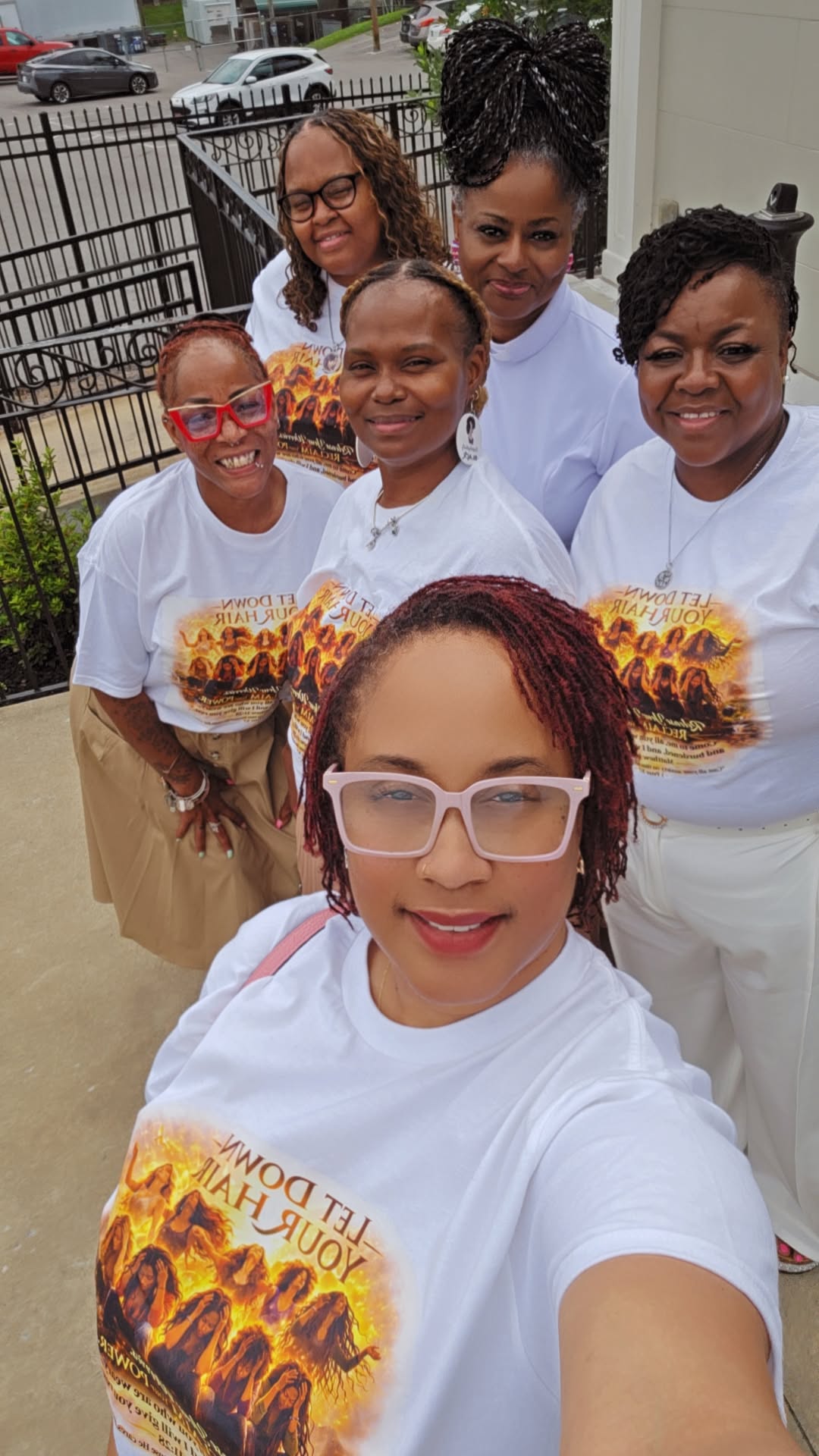 Group of six women standing outdoors, smiling, wearing matching white T-shirts with a colorful design, in front of a fence and buildings.