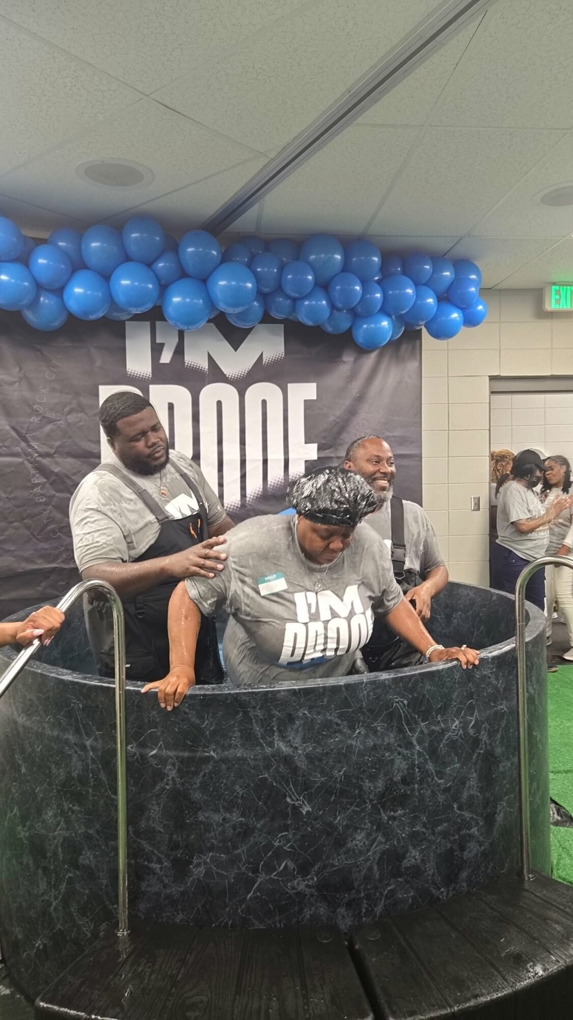 People standing behind a black marble baptismal font with blue balloons above, in a room with a black backdrop and a green exit sign.