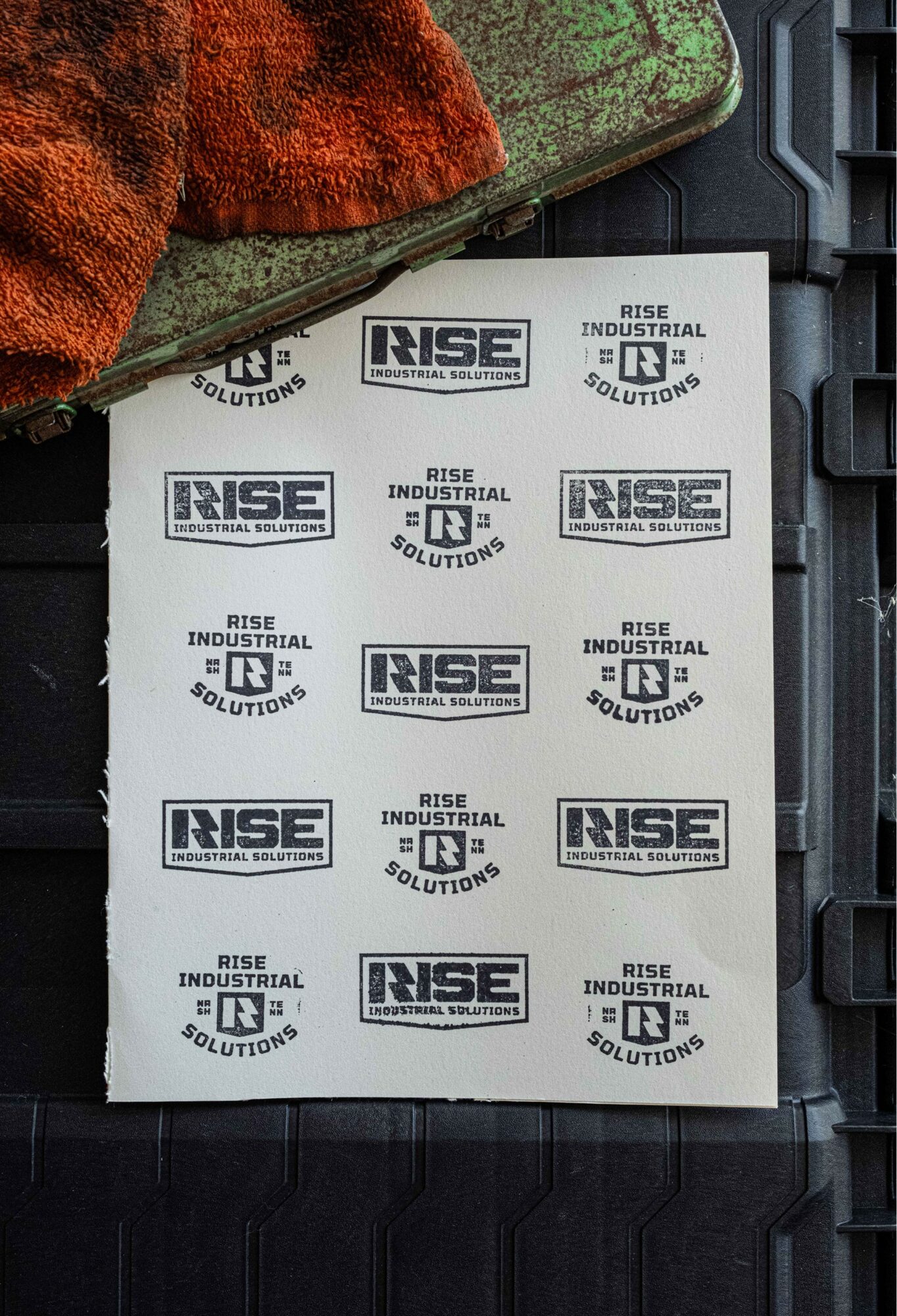 Stack of stickers with 'RISE INDUSTRIAL SOLUTIONS' logo on a black surface, partially covered by a textured fabric.