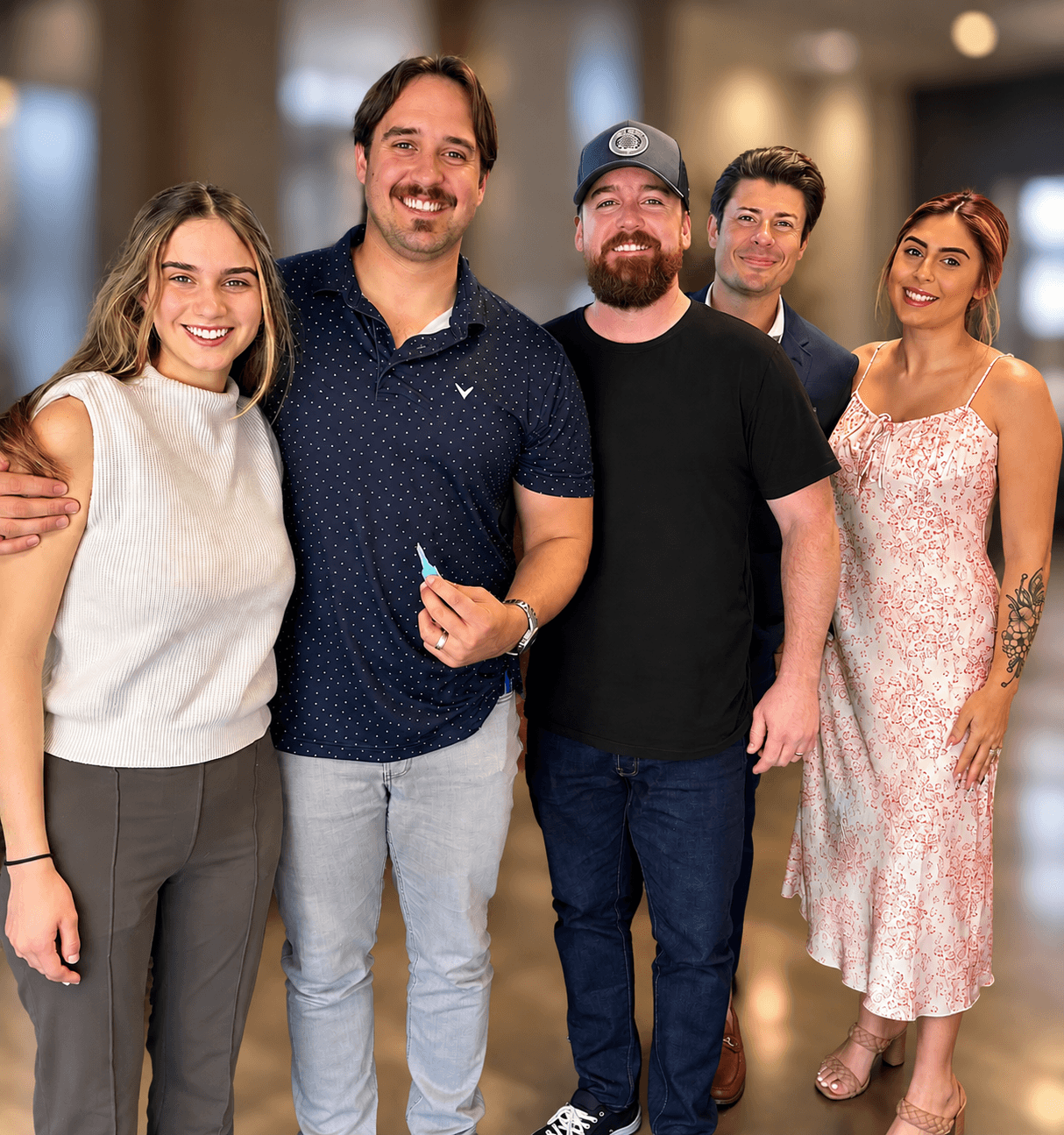 Group of five smiling people standing indoors, posing for a photo, with blurred background. Word count: 20.