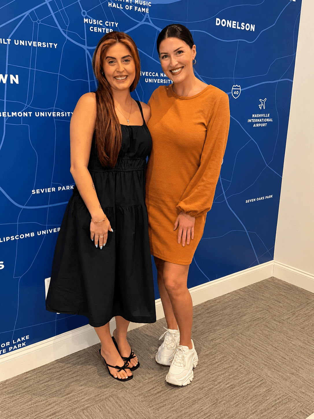 Two women standing in front of a blue map background, smiling, one in a black dress and the other in an orange dress, both wearing sneakers.