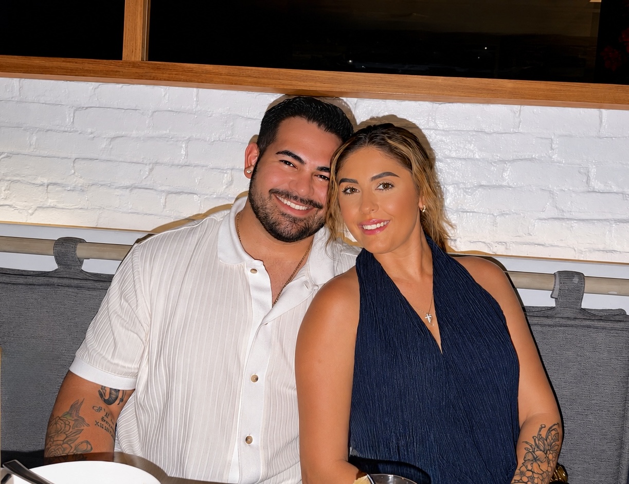 A smiling man and woman sit close together indoors against a white brick wall, posing for a photo.