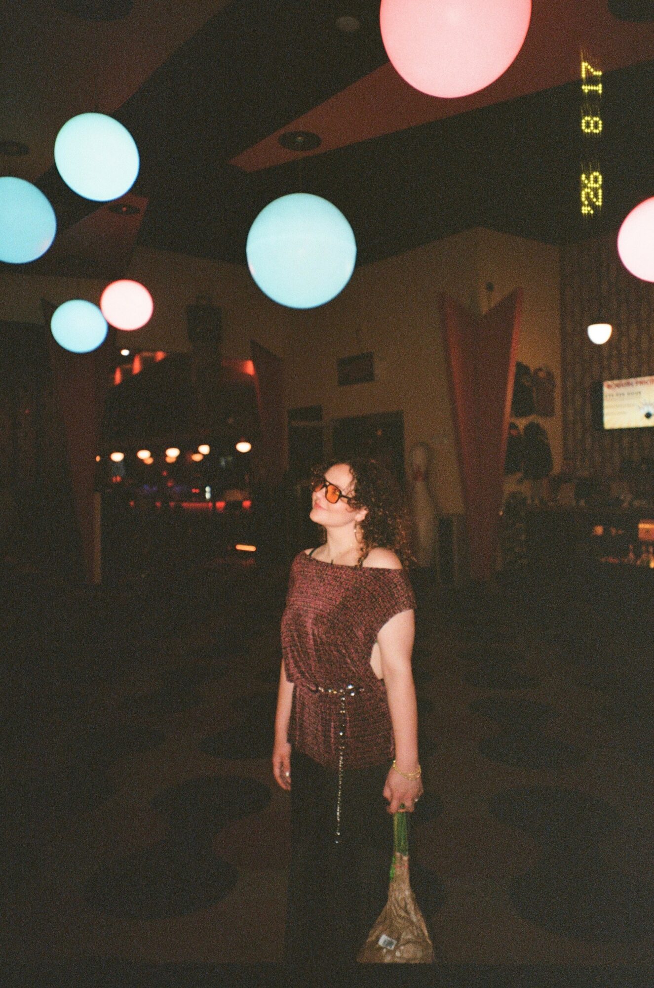 Woman in a dark dress with glasses standing in a dimly lit space with colorful hanging lights.