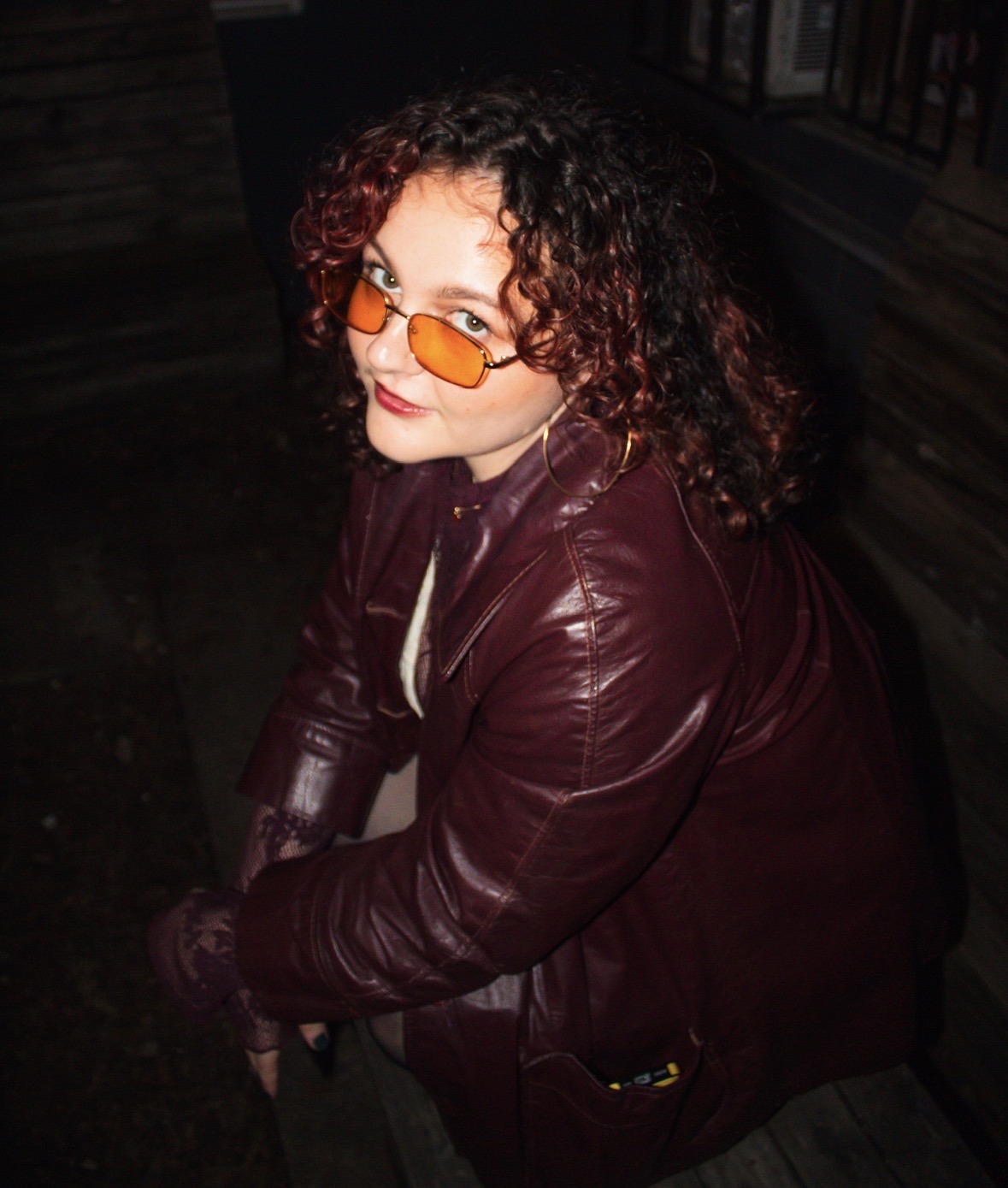Person with curly hair wearing orange-tinted glasses and a maroon leather jacket, sitting outdoors at night.