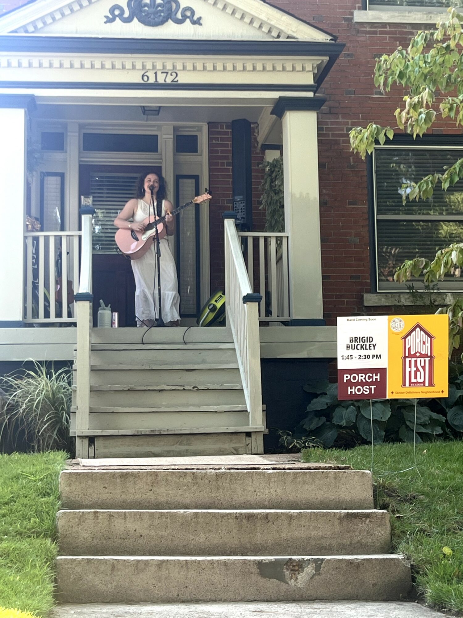 Person with guitar standing on porch steps of a brick house, holding a microphone, with a sign nearby, daytime.