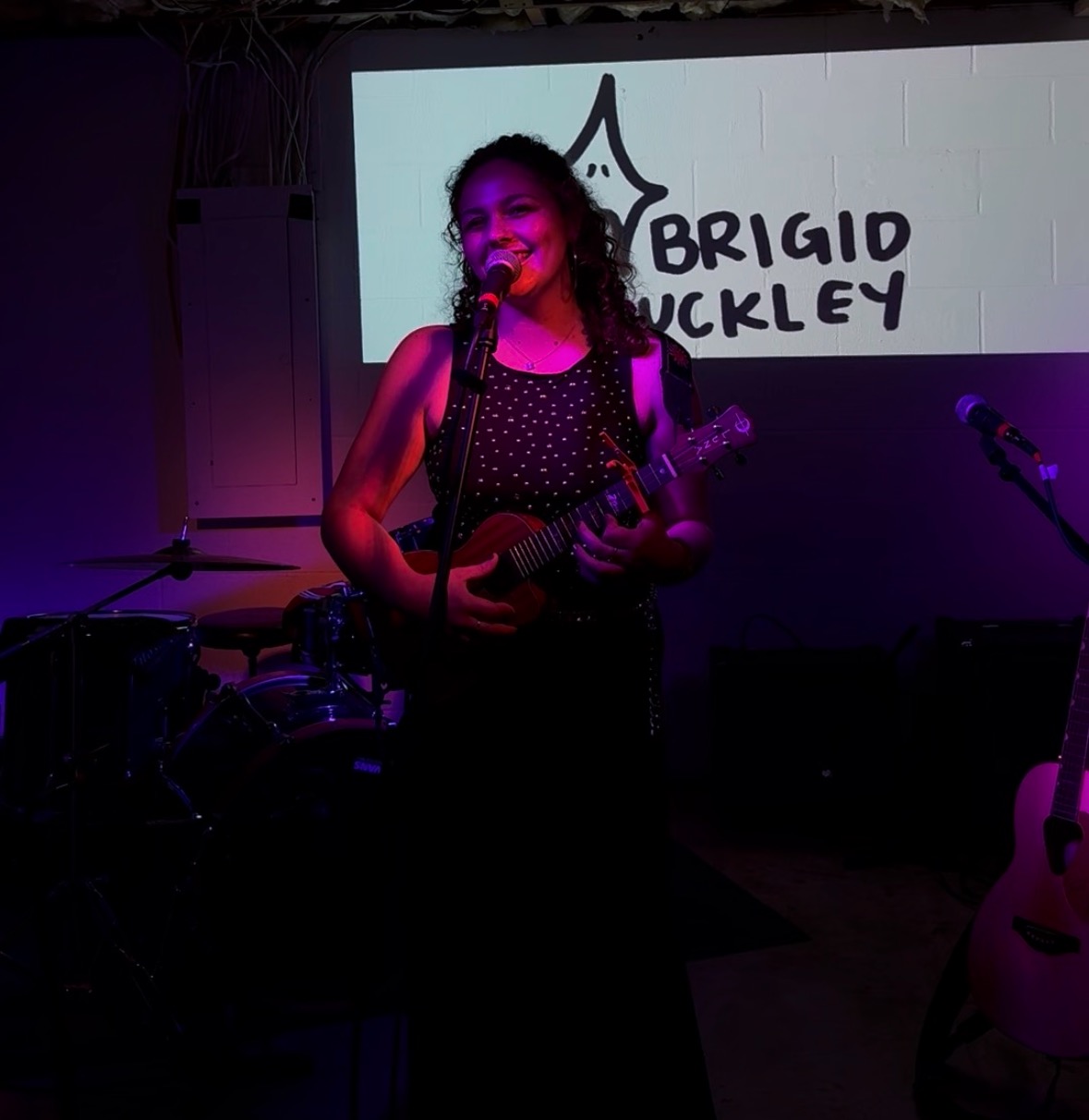 Young woman singing and playing ukulele on stage with a microphone, background screen displaying text, dim lighting.