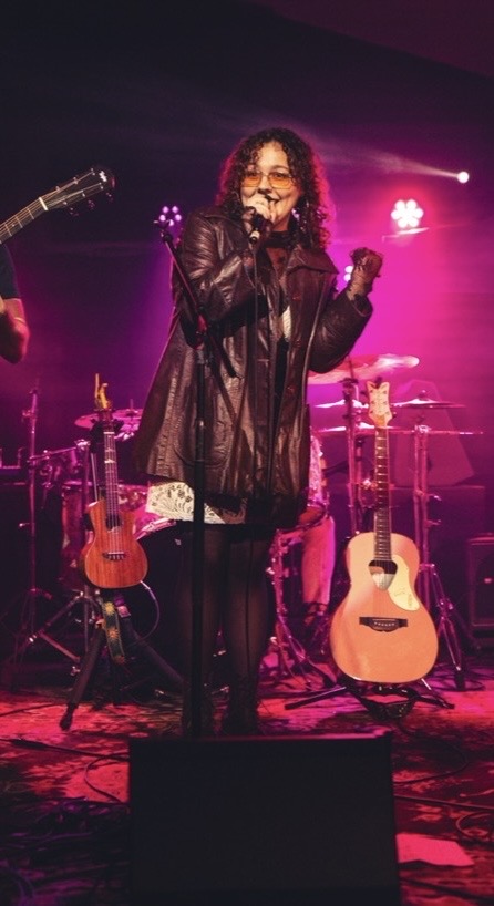 A woman with curly hair and glasses sings into a microphone on stage, surrounded by guitars and pink stage lights.