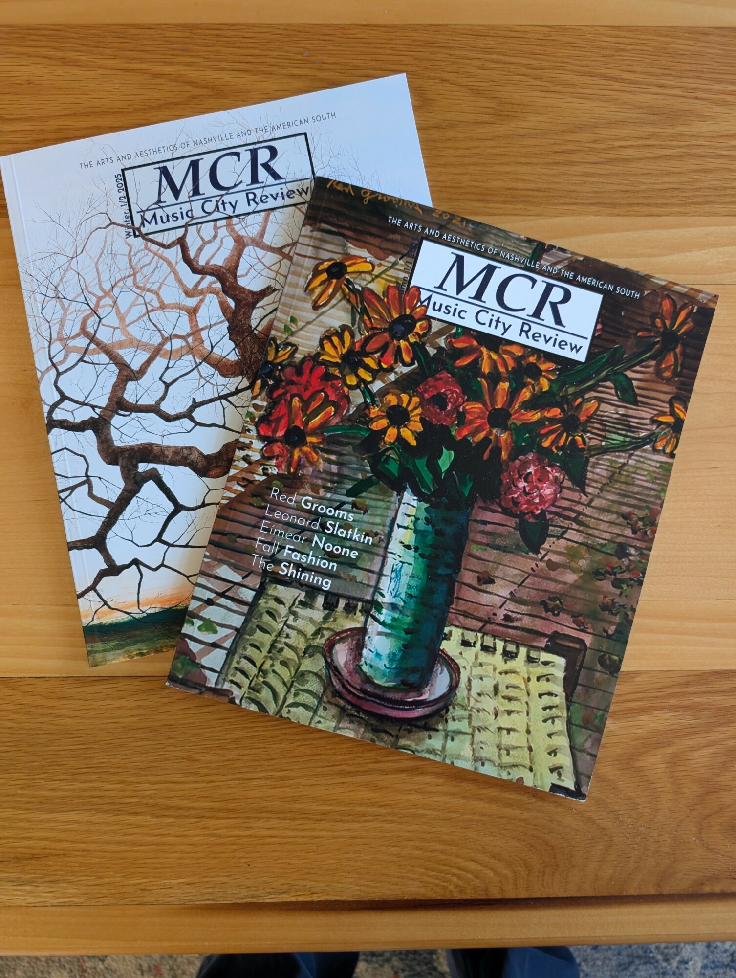 Two magazines titled 'Music City Review' on a wooden surface, one with a tree branch illustration and the other with a colorful vase and flowers.