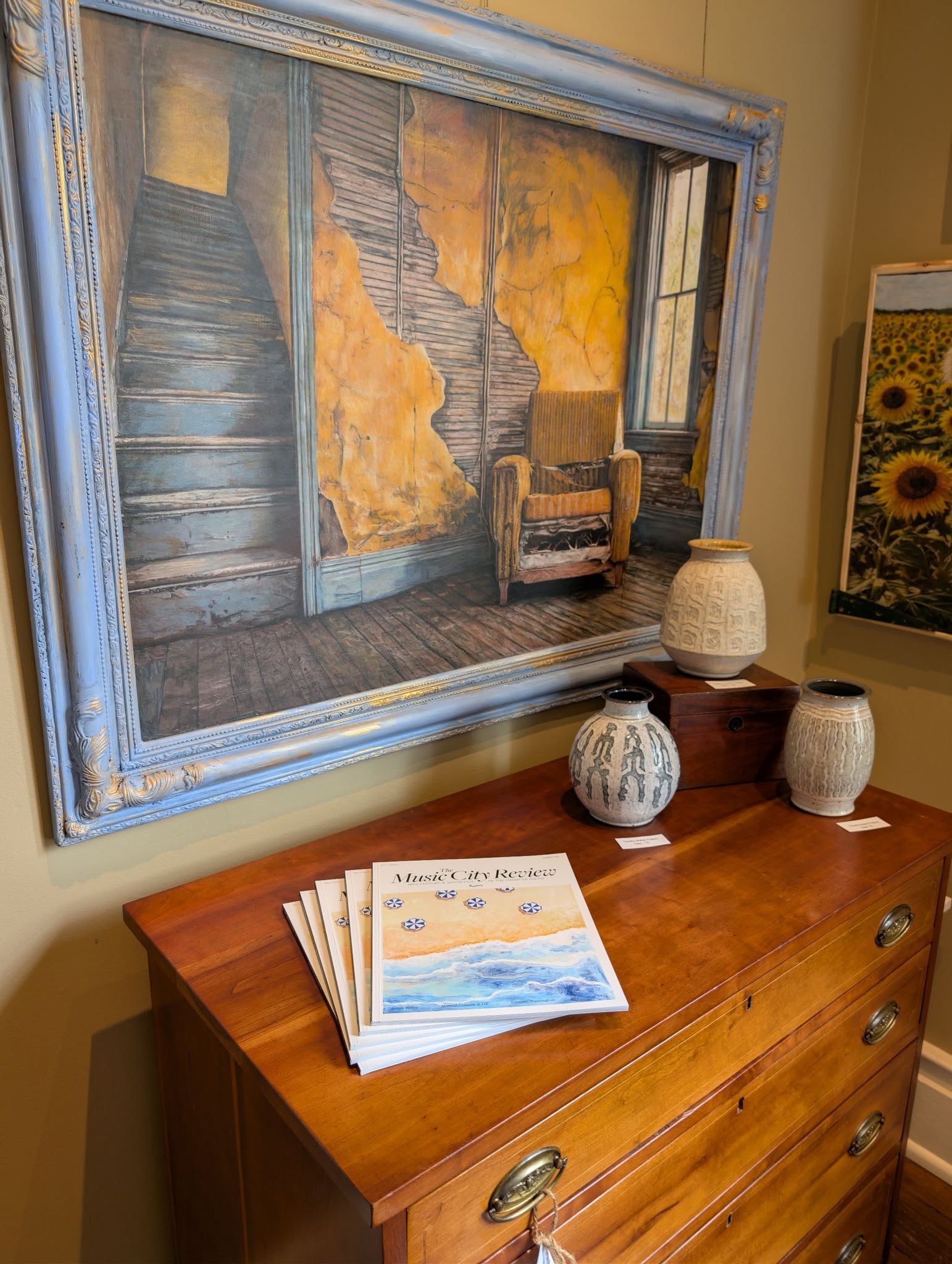 Painting of stairs and a window hangs above a wooden dresser with books and decorative vases.