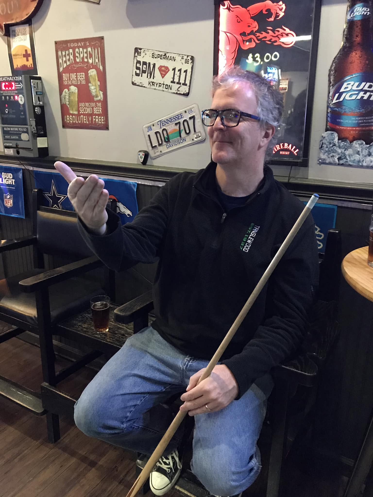 Man sitting in a bar holding a pool cue, wearing glasses and a black jacket, with posters on the wall behind him.