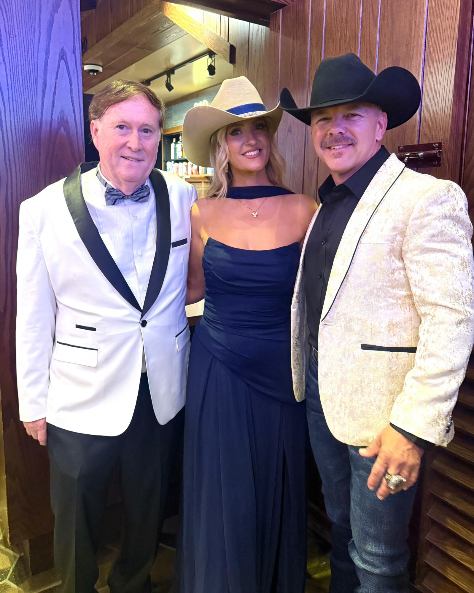 Three people dressed in formal and western attire standing indoors, smiling at the camera.