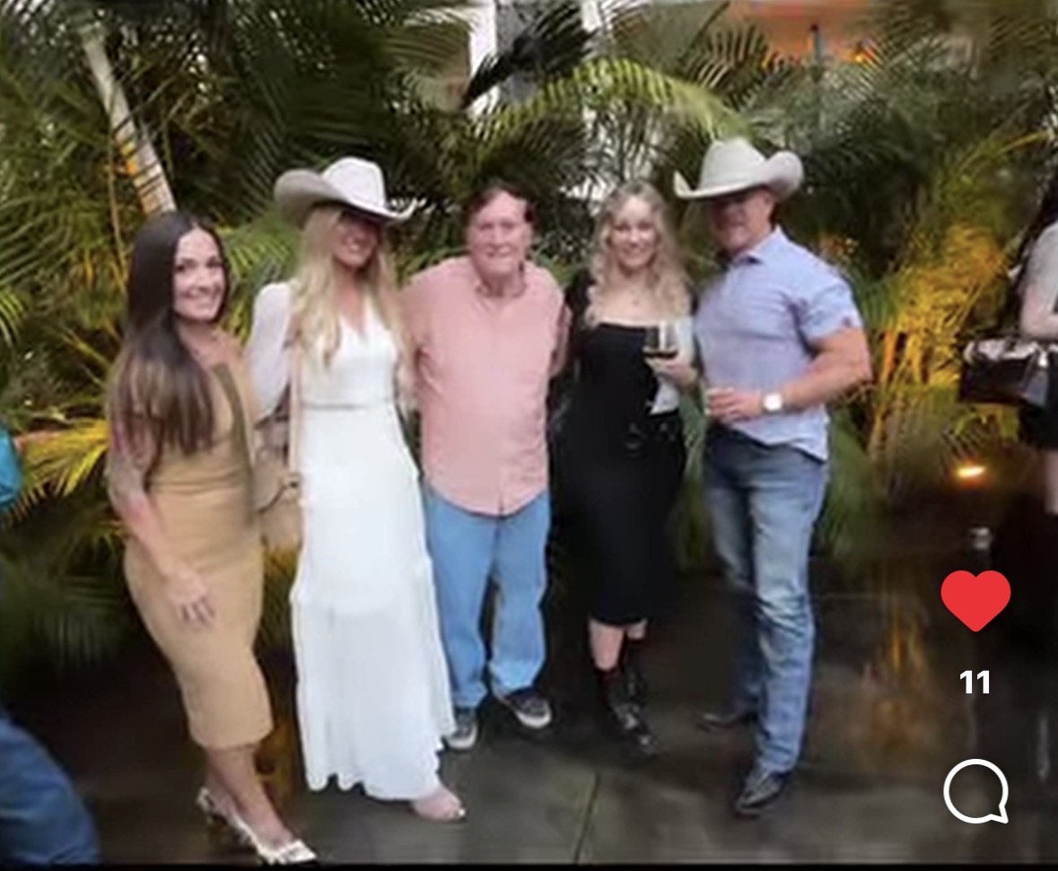 Five people standing outdoors with plants, smiling, some wearing hats, holding drinks, in a social setting.
