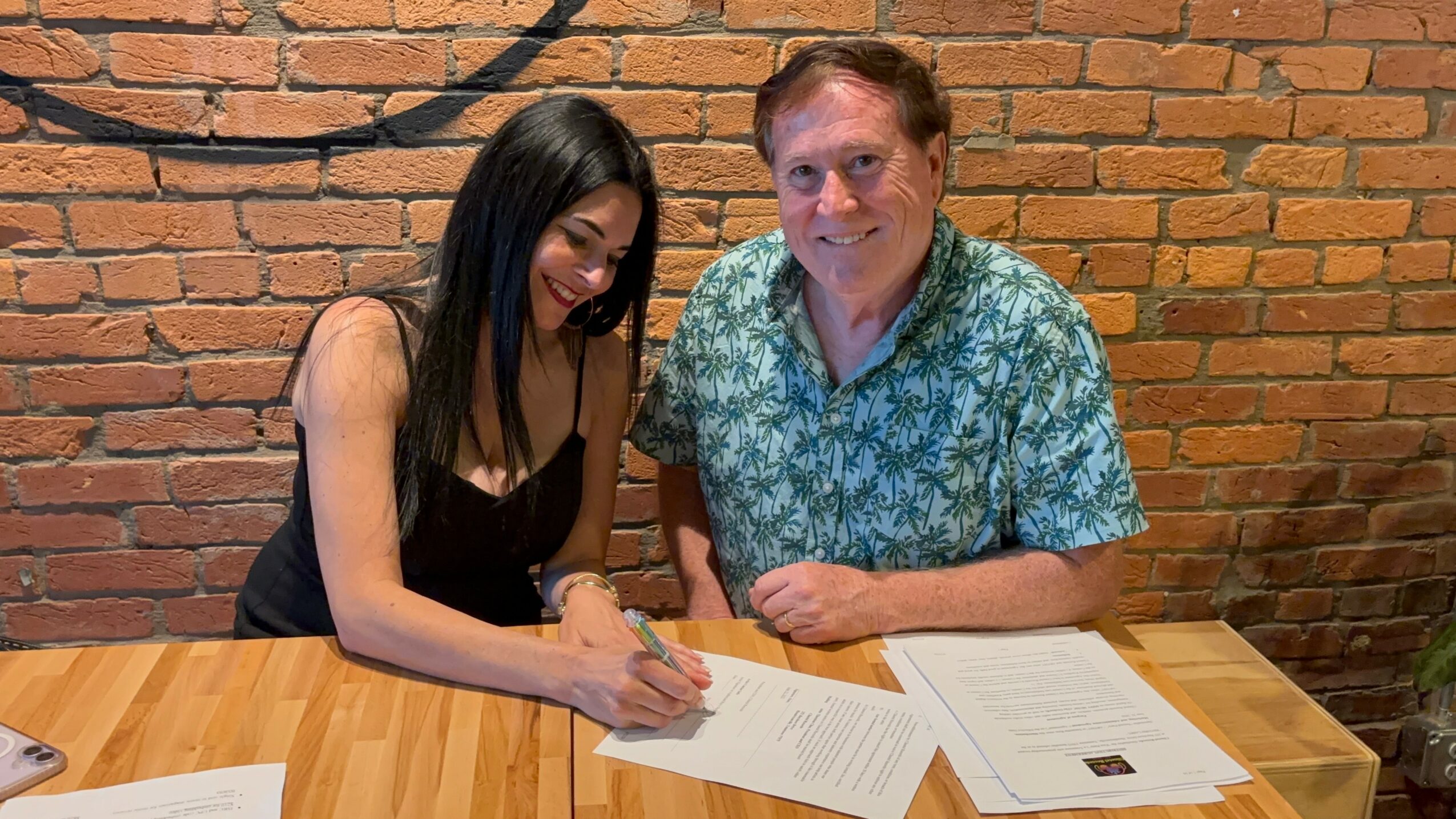 A woman and a man sit at a wooden table signing documents against a brick wall background.