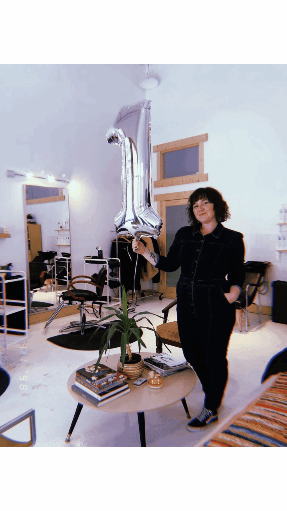Woman standing in a room with a large silver balloon shaped like the number 1, plants, books, and furniture.