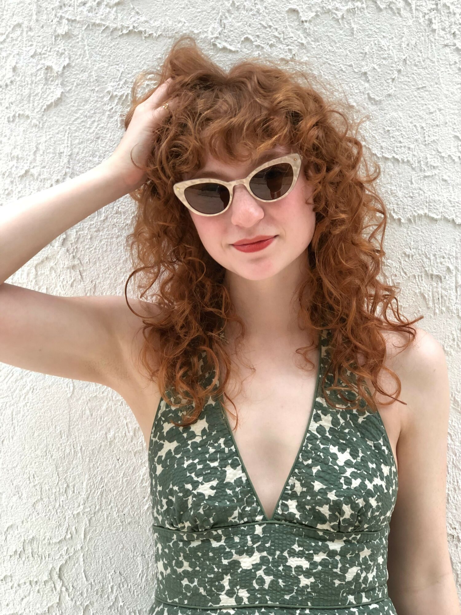Woman with curly red hair wearing sunglasses and a patterned dress, leaning against a textured white wall.