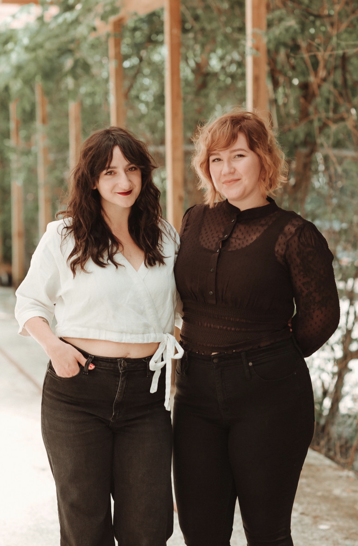 Two women standing outdoors, one in a white top and black pants, the other in a black lace top and black pants, smiling.