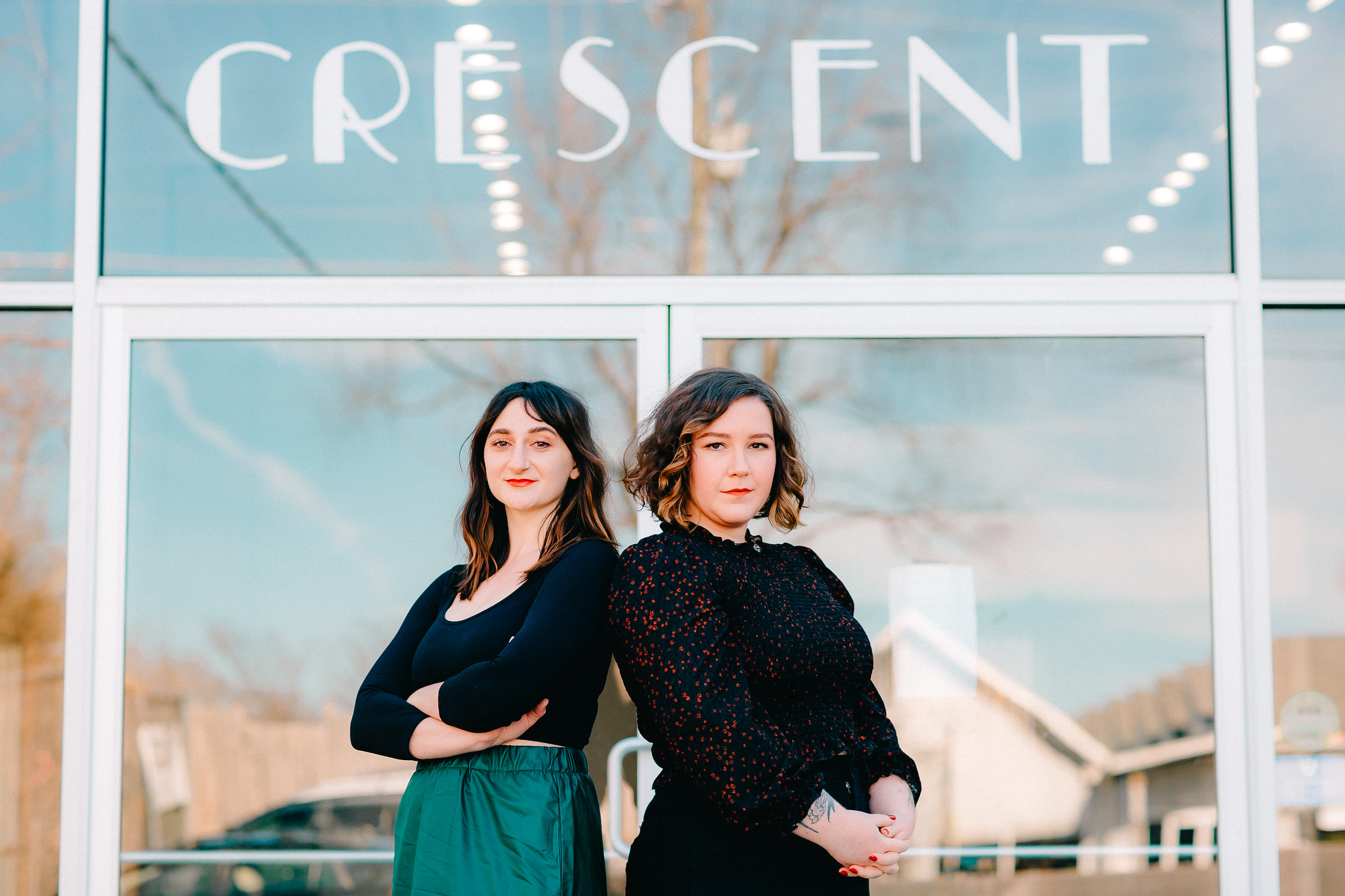 Two women stand in front of a glass window with the word 'CRESCENT' written above, reflecting a cloudy sky.