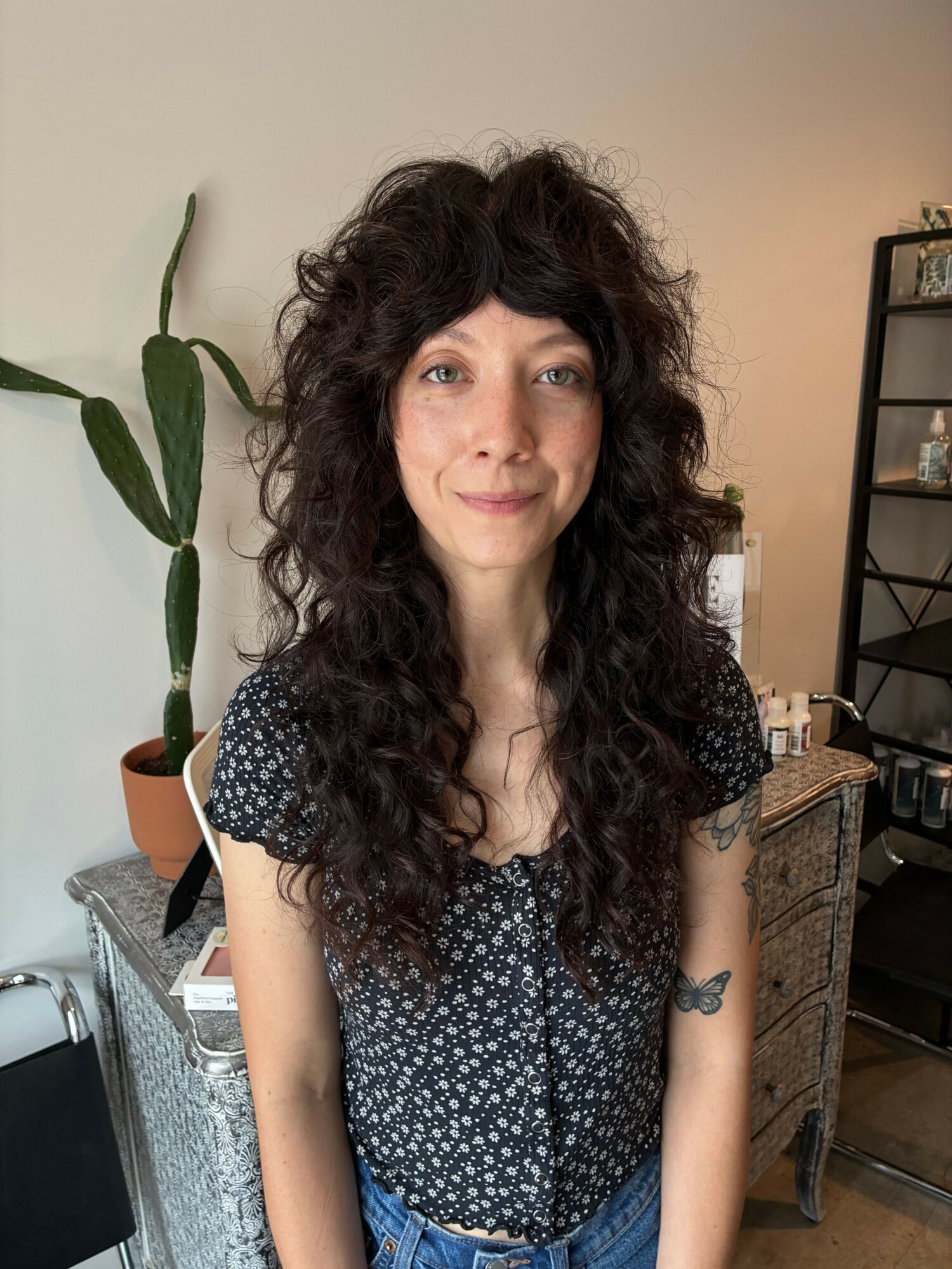 Woman with long curly hair wearing a dark patterned top, standing indoors with a plant and furniture in the background.