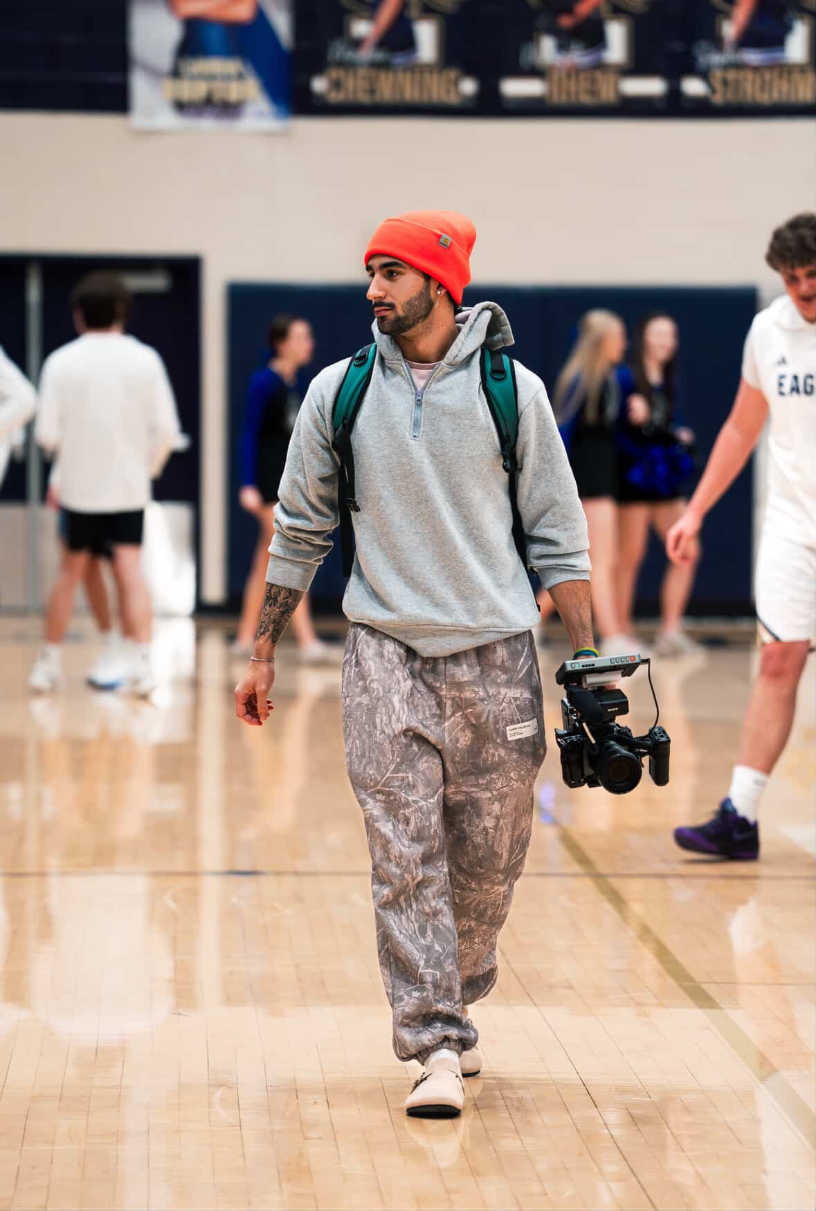 Young man walking in a gymnasium with a camera, wearing a red beanie, gray hoodie, patterned pants, and a backpack.