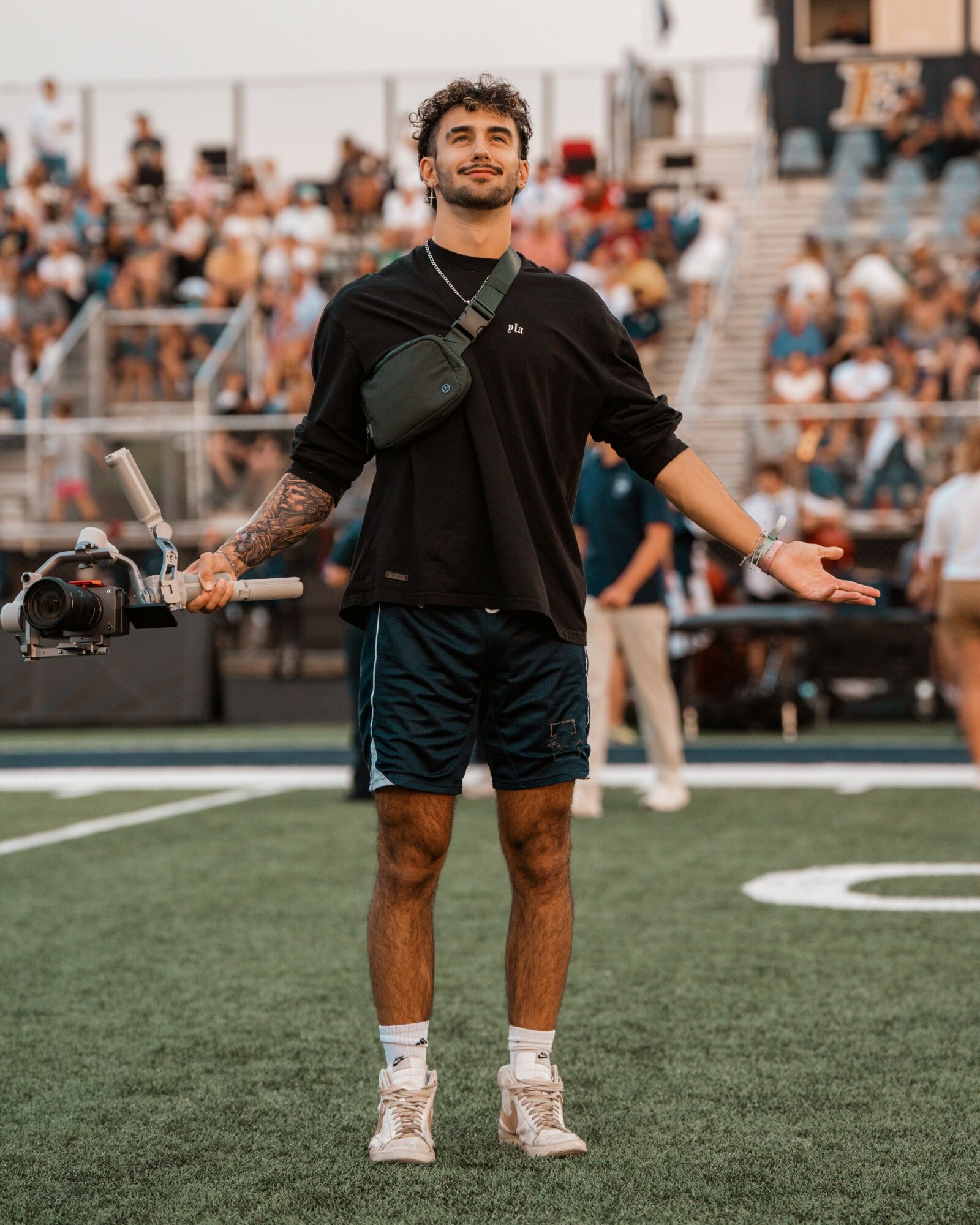 Young man standing on a sports field with arms outstretched, holding a camera stabilizer, surrounded by spectators.