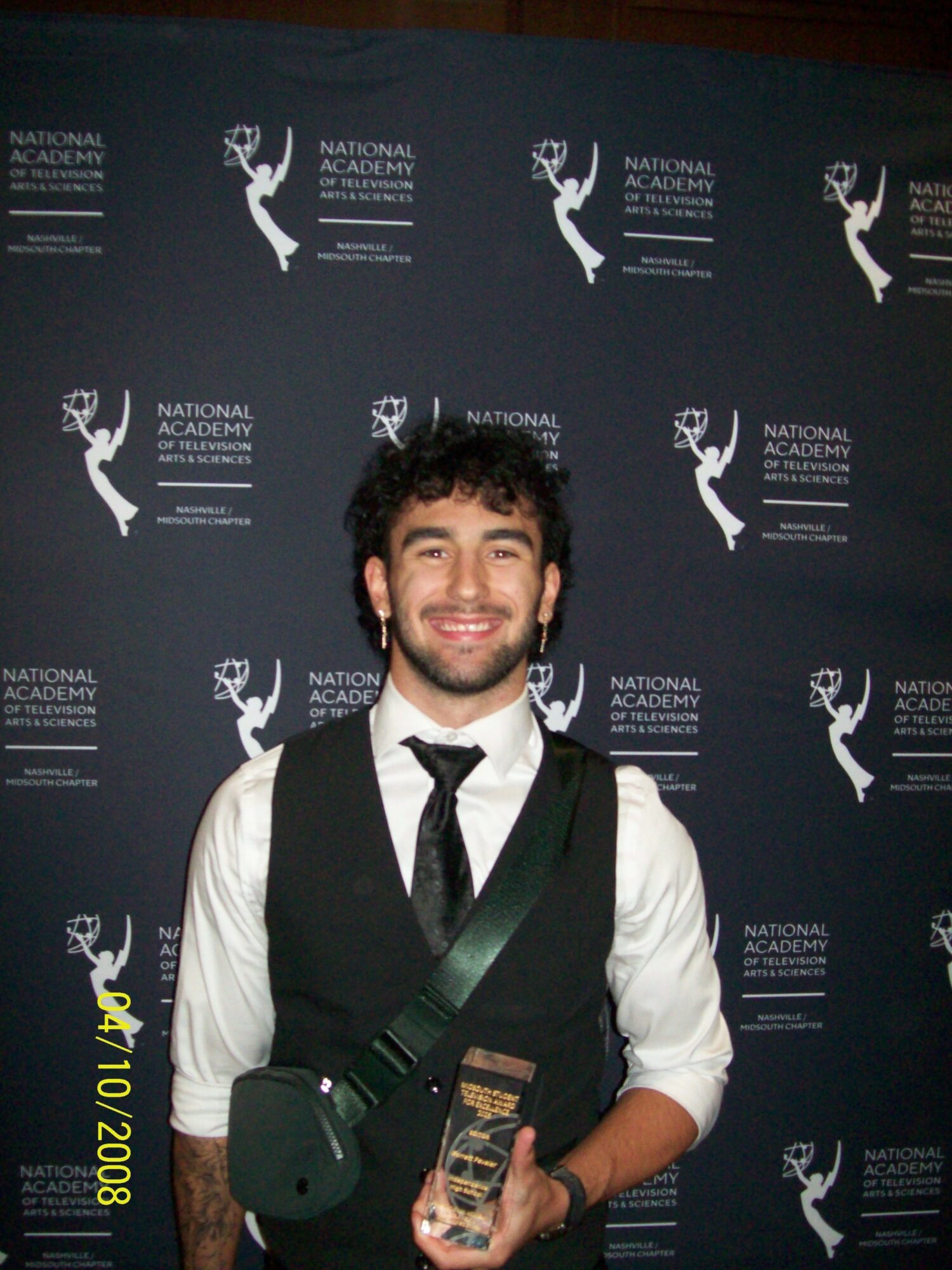 Young man with dark curly hair and beard, smiling, holding an award, standing in front of a backdrop with logos.