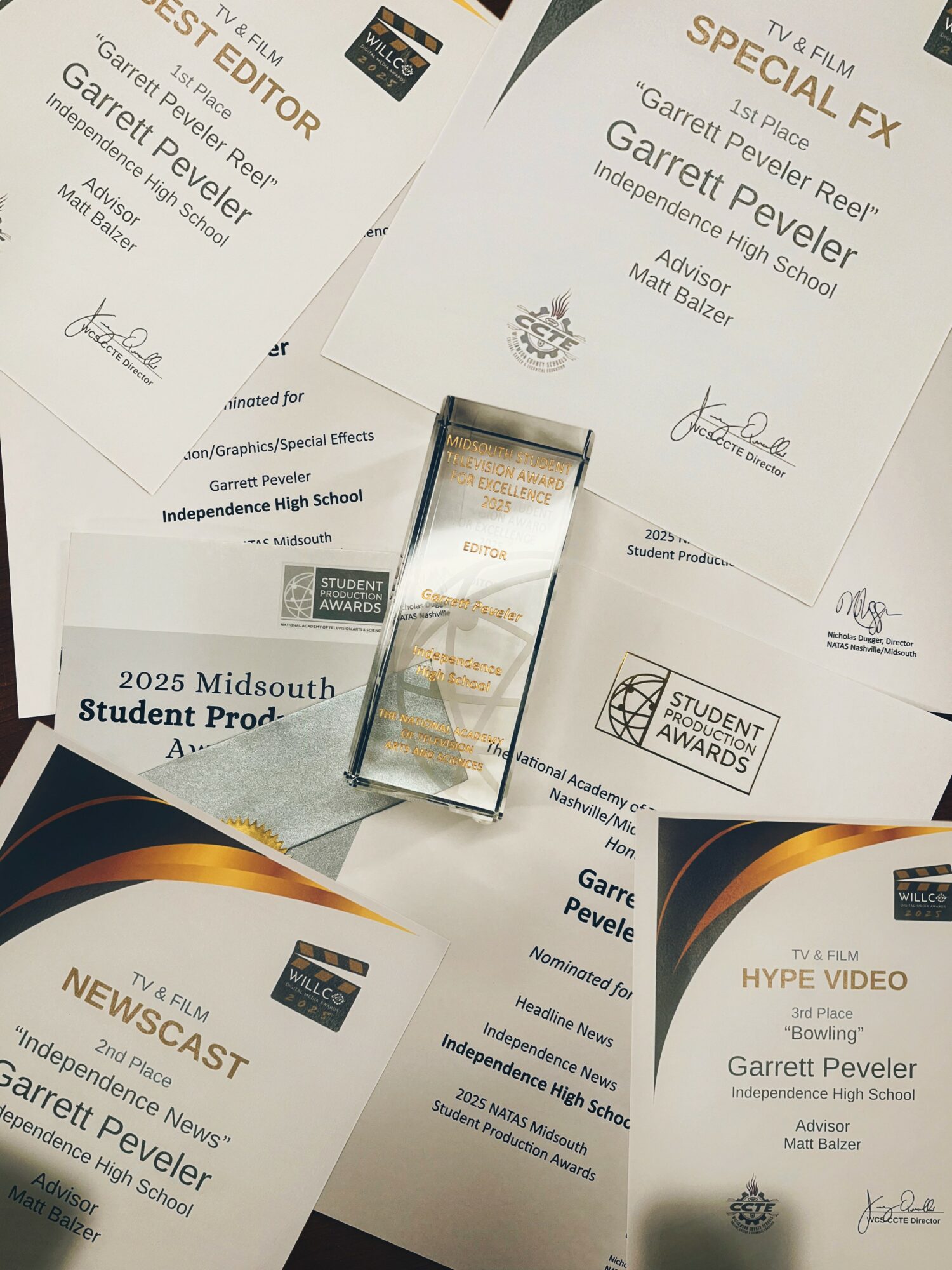 Multiple awards and certificates for Garrett Peveler displayed on a table, some overlapping, with a small glass award in the center.
