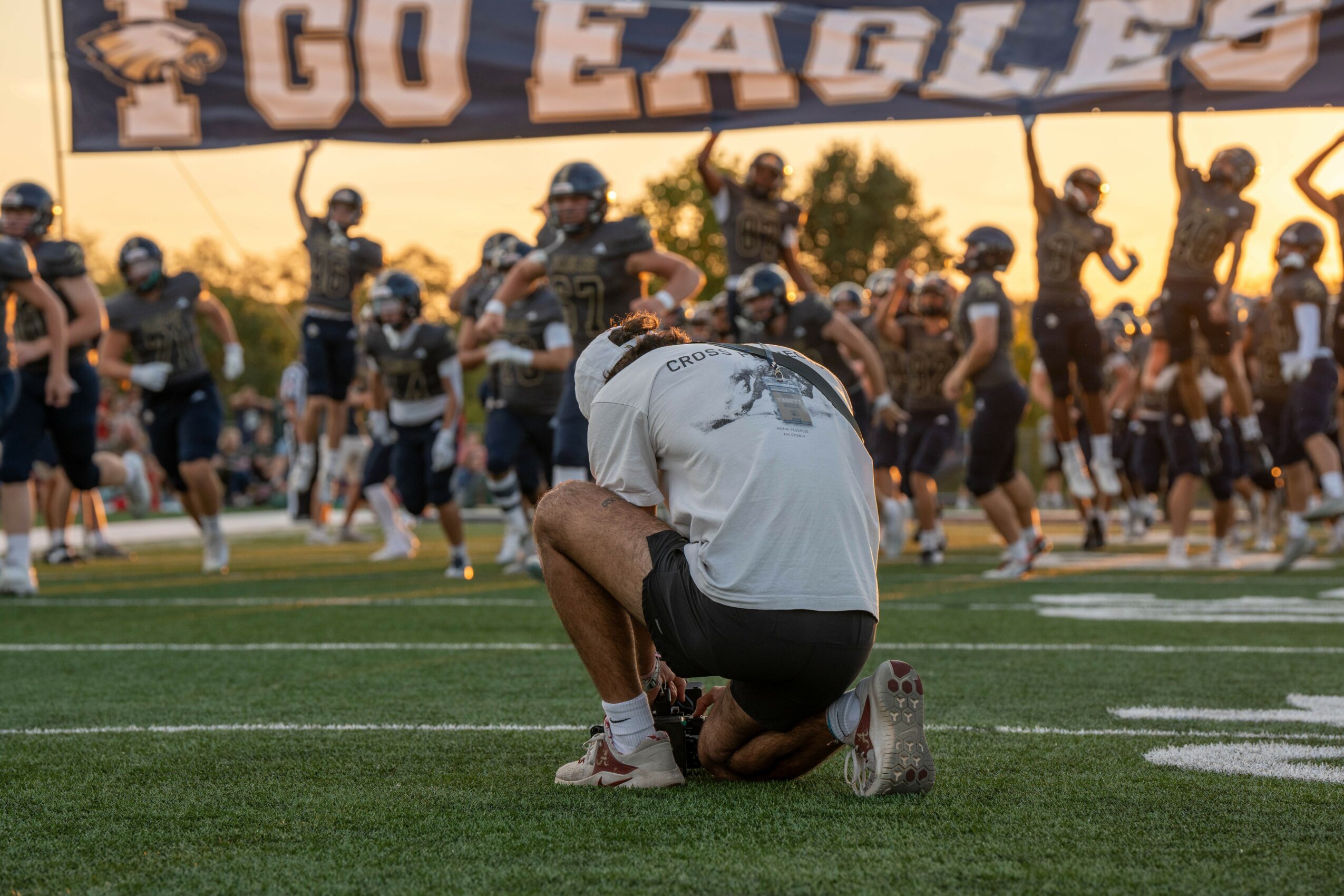 Football team on field with a person kneeling in foreground, players in background, sunset sky, large banner, grass field