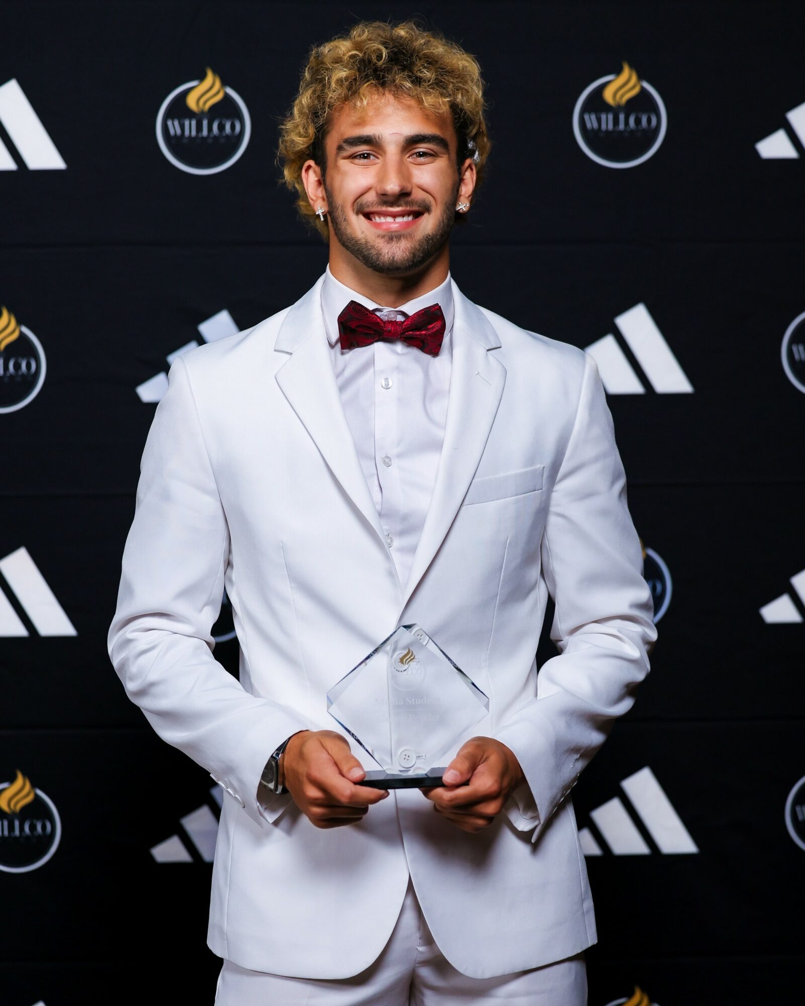 Young man in a white suit and red bow tie holding an award, smiling, standing in front of a black background with logos.
