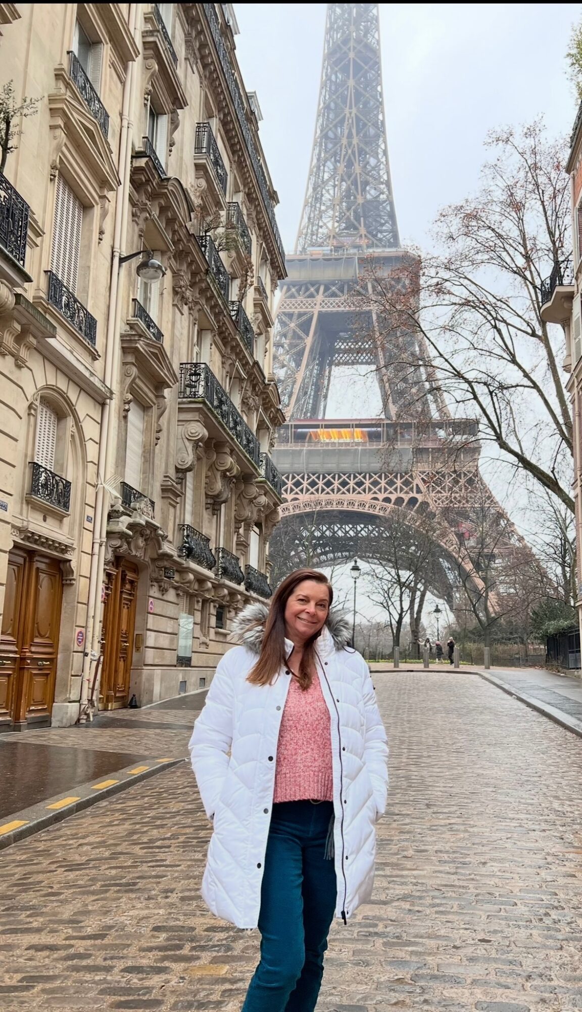 Woman standing on cobblestone street with Parisian buildings and Eiffel Tower in background.