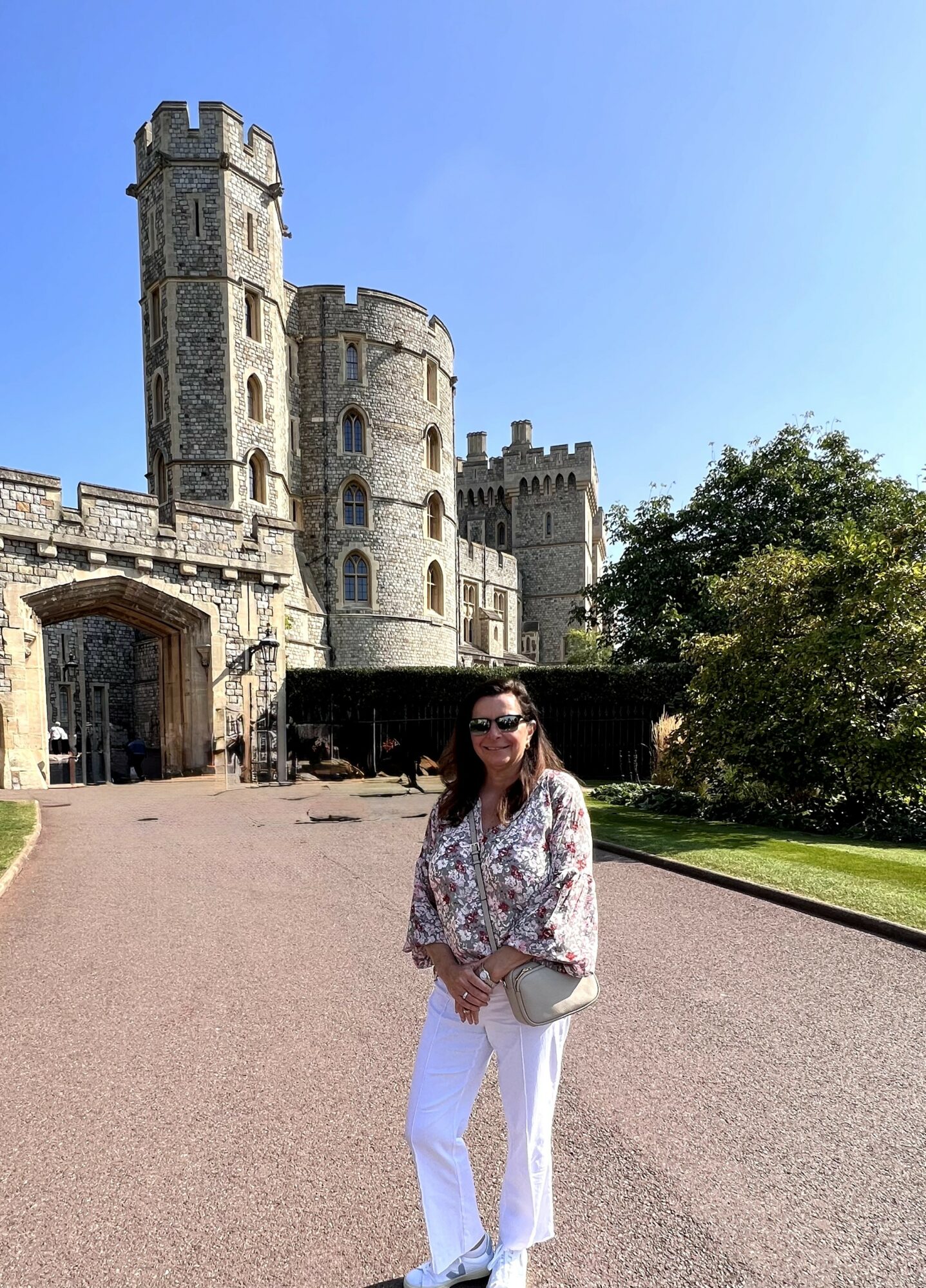 Woman standing in front of a castle with tall towers, trees, and a clear blue sky.