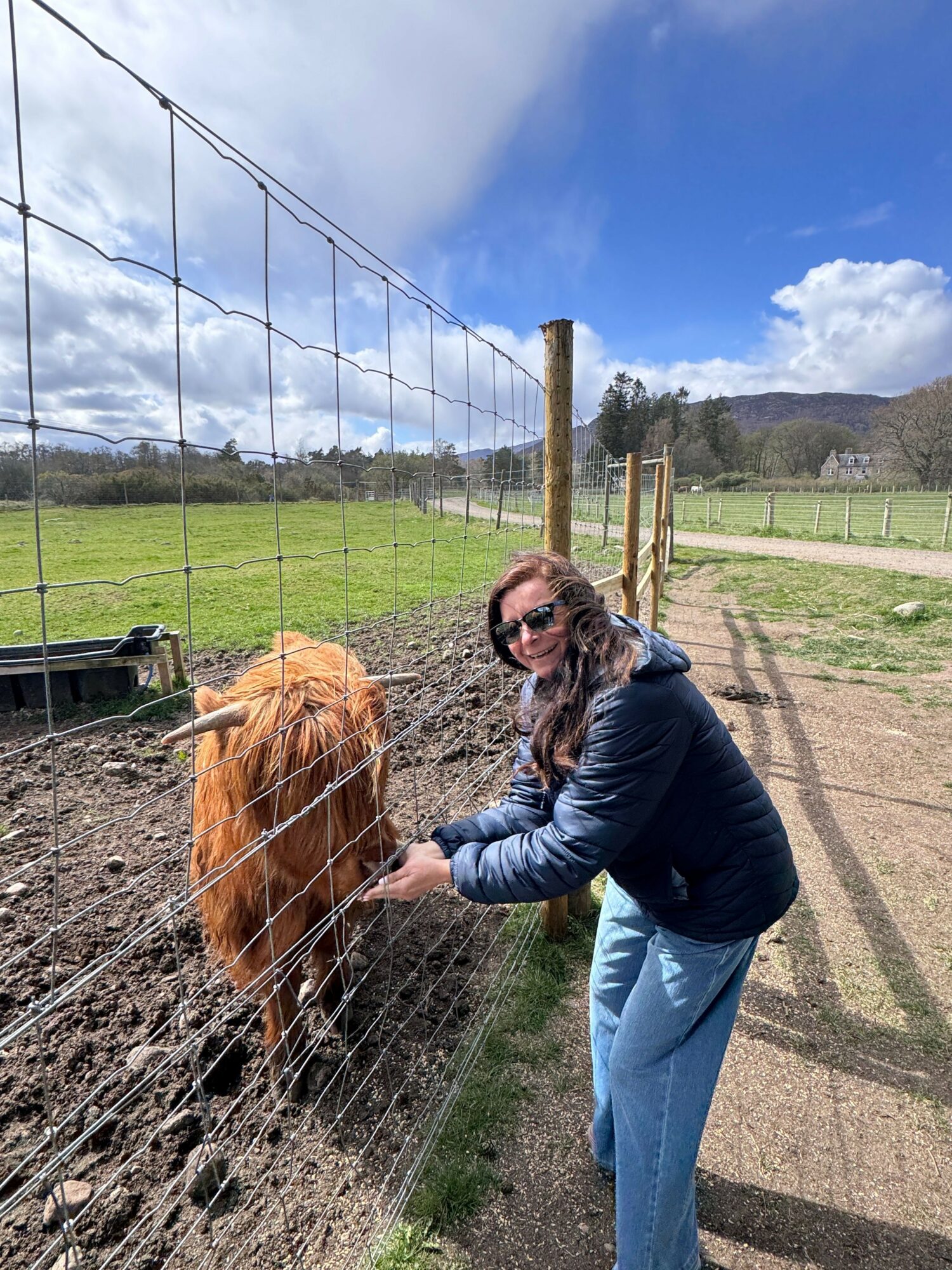Person with long hair and sunglasses leaning over a wire fence, interacting with a dog in a grassy outdoor area.