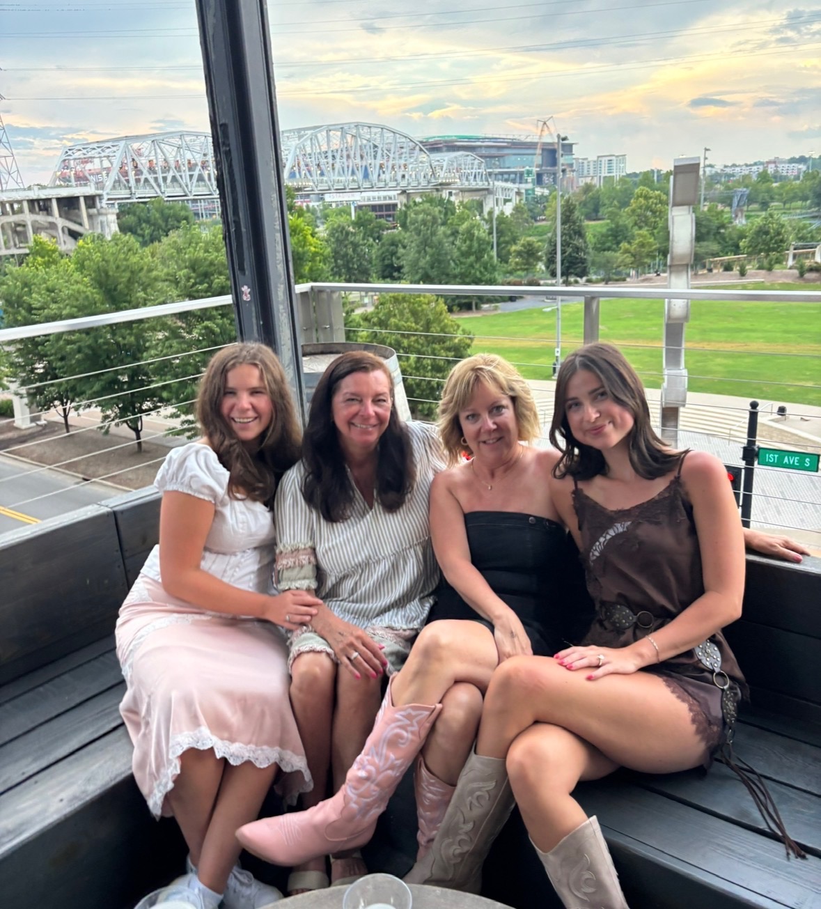 Four women sitting together on a bench inside a glass-walled structure with a cityscape and bridge in the background.