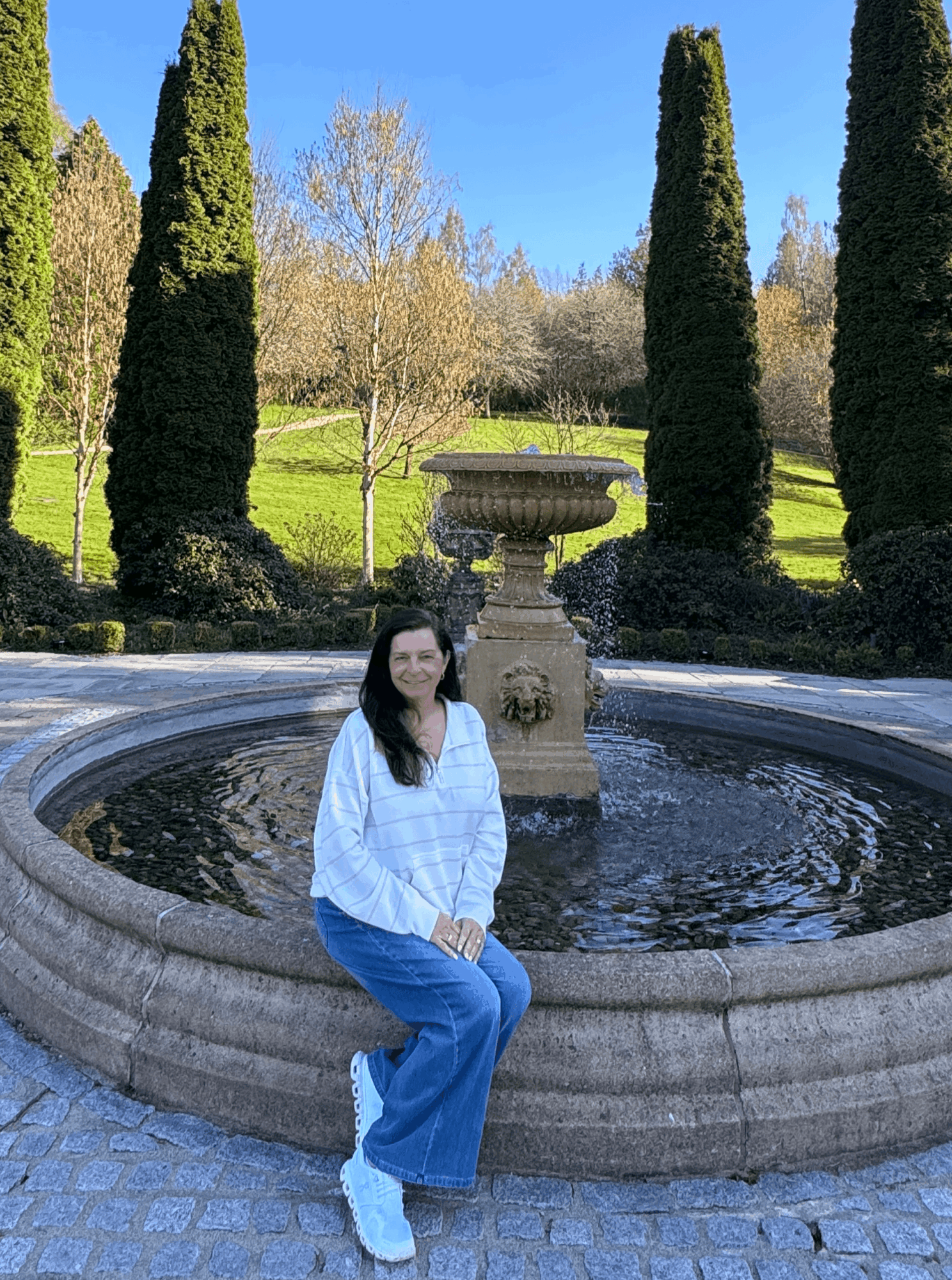 Woman sitting on fountain edge in a park with tall trees and a clear blue sky.