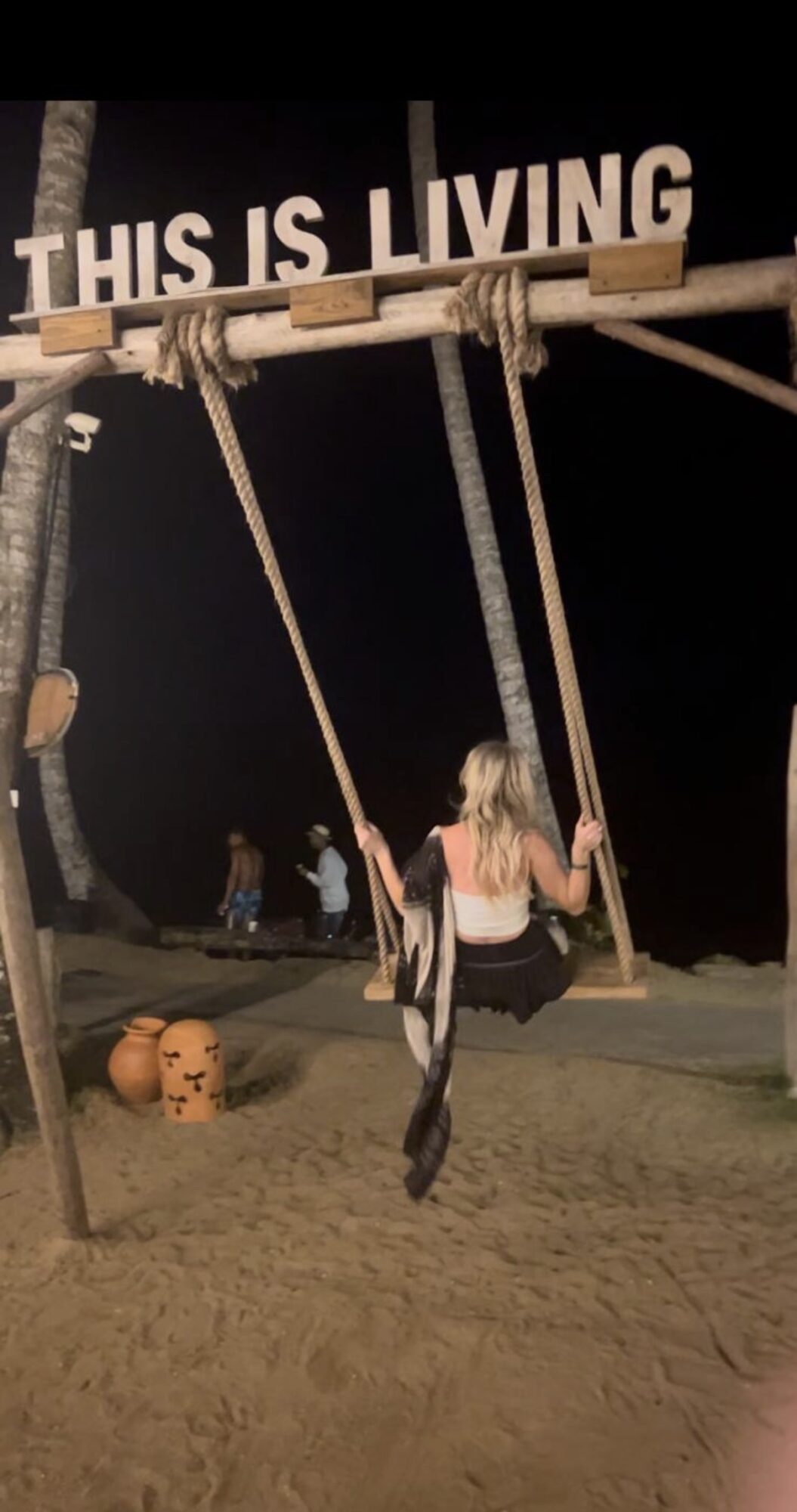 Person sitting on a swing at night with a sign reading 'THIS IS LIVING' above, and pumpkins on the ground nearby.