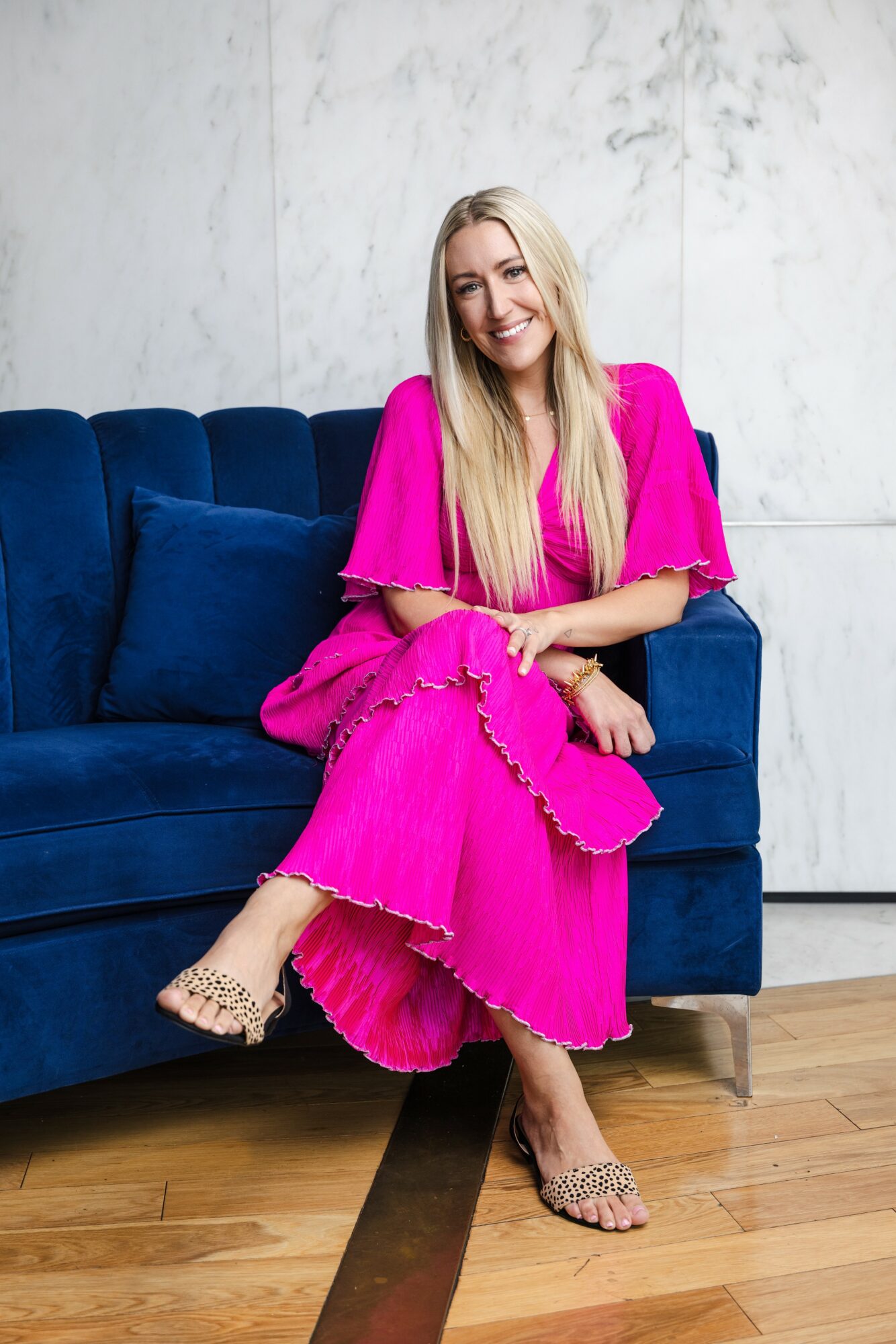 Woman with long blonde hair in a bright pink dress sitting on a blue sofa, smiling, against a white wall.