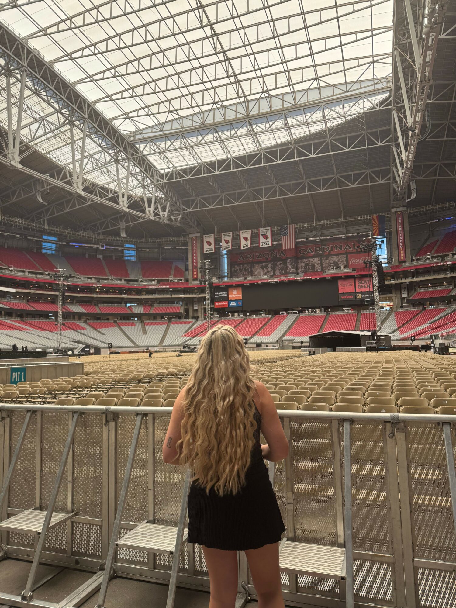 Person with long blonde hair standing in an empty stadium with red seats and a large glass ceiling.