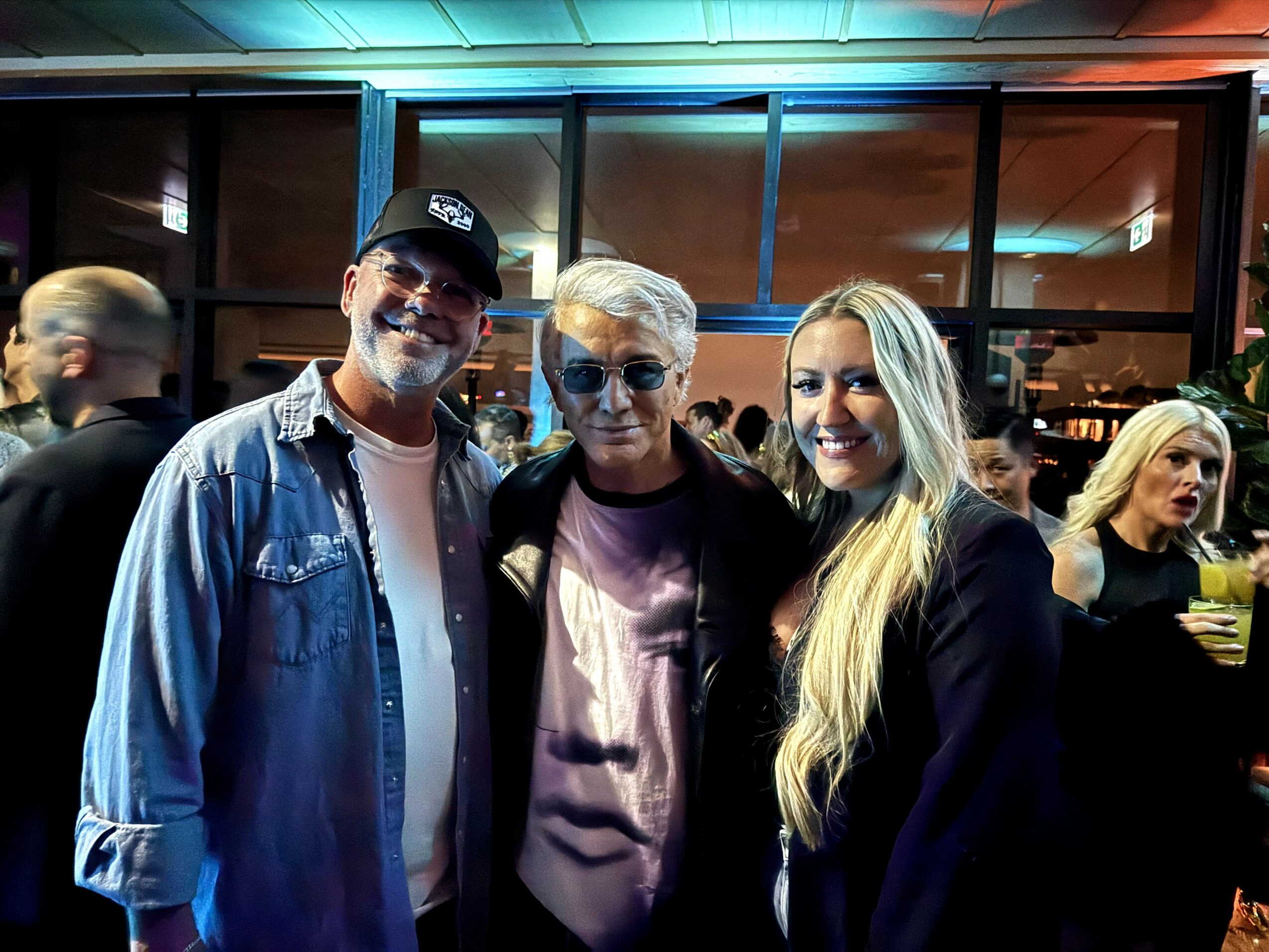 Three people standing together indoors, smiling, with a crowd in the background.