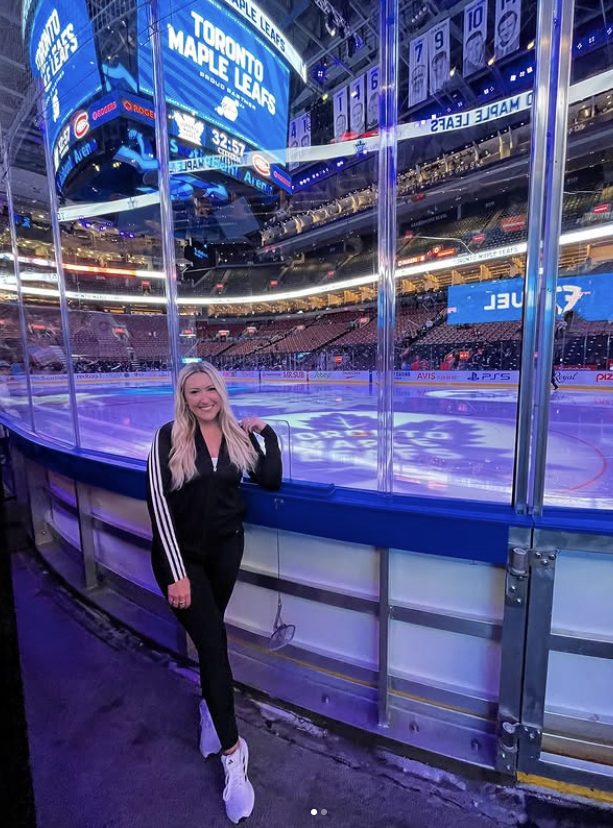 Person with long blonde hair standing near ice rink glass at a hockey arena, illuminated by bright digital screens.