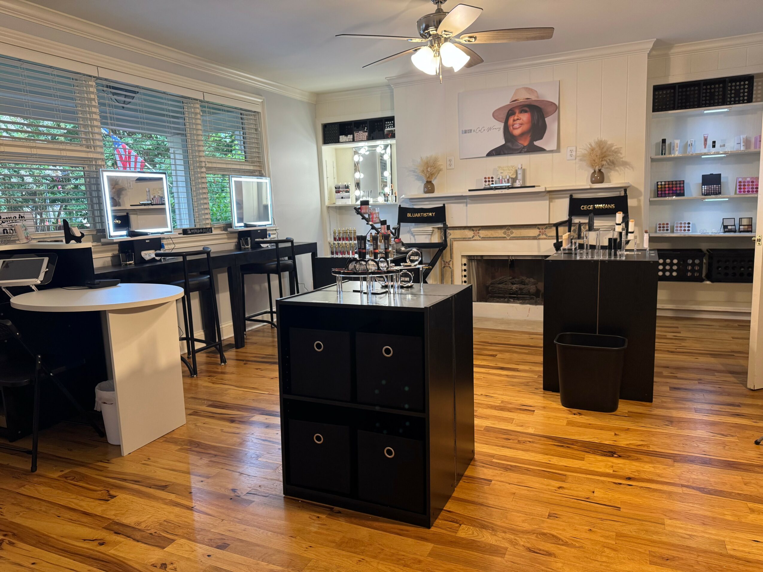 Room with three computer desks, a central display table, a fireplace, and shelves with products, bright windows, and wooden floor.