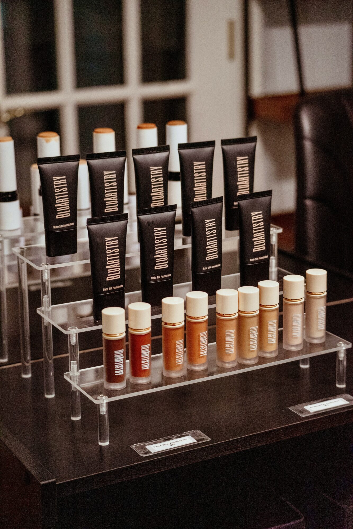 Display of skincare or cosmetic products in black tubes and small bottles on a clear acrylic stand, on a dark table.